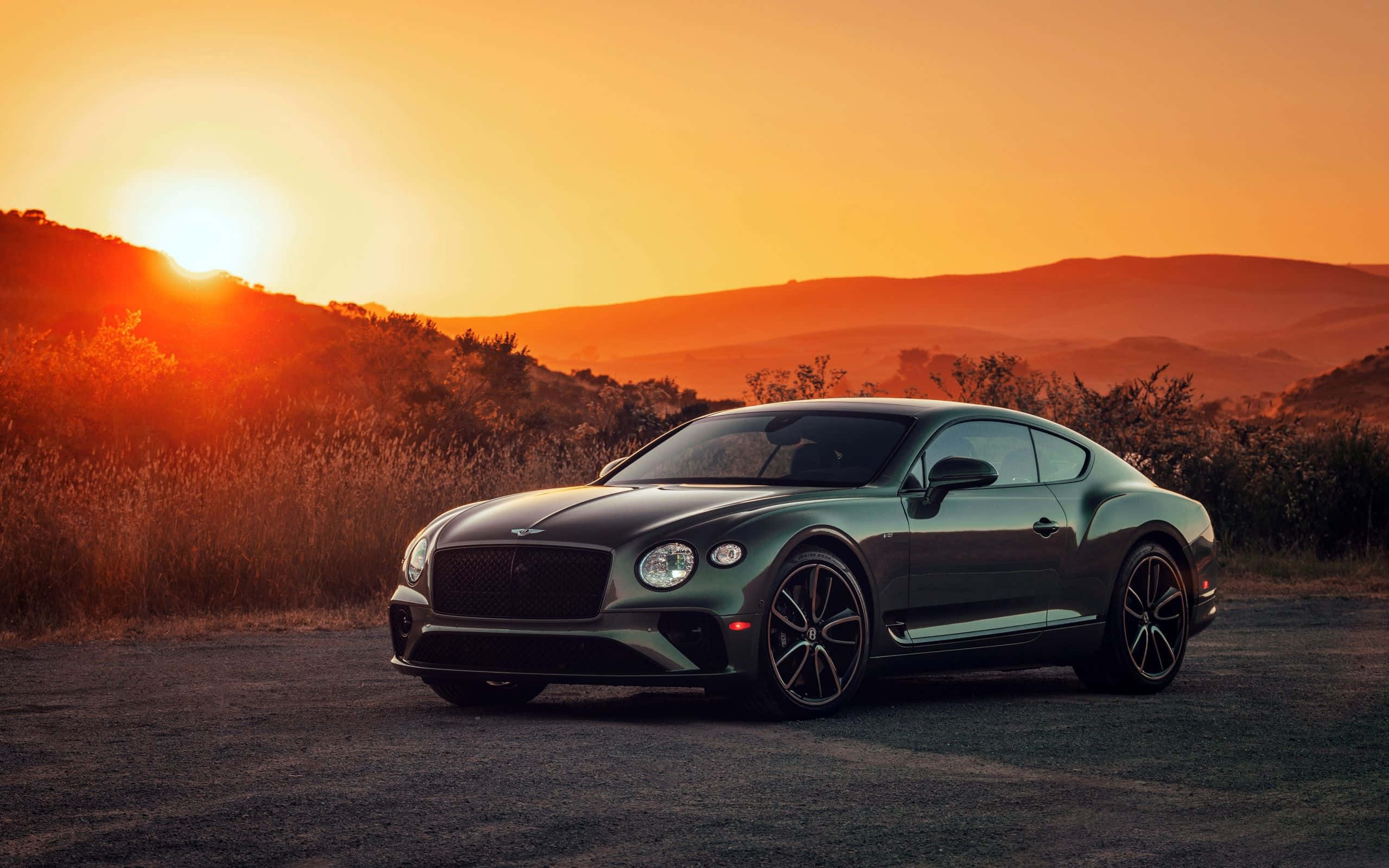 奢華 Bentley 駛上街道 壁紙 — 一架時尚的綠色賓利 Continental GT 停泊在日落時分的泥路上，背景是沐浴在金色光芒中的起伏山丘。