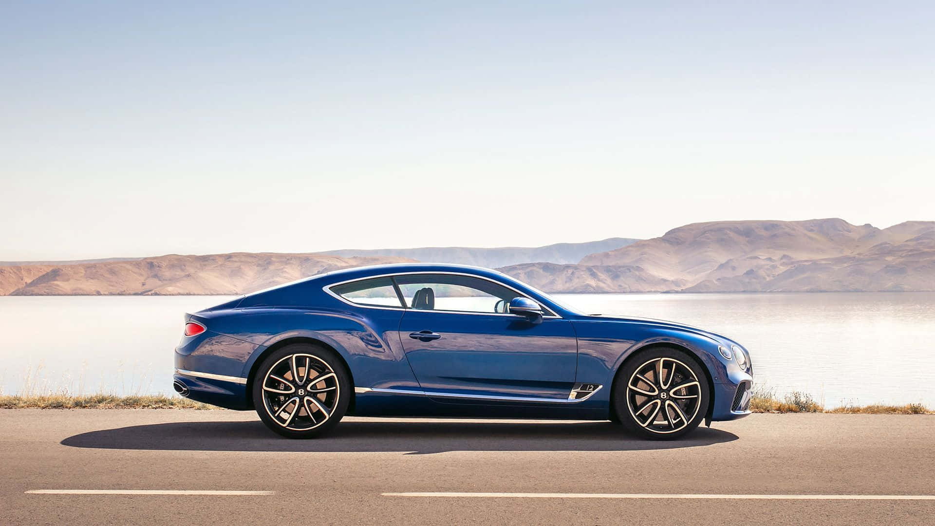 奢華的巔峰 — Bentley 壁紙 — 一架時尚的藍色賓利 Continental GT 雙門跑車停泊在路邊，俯瞰著平靜的湖泊和遠處的山脈，天空晴朗。