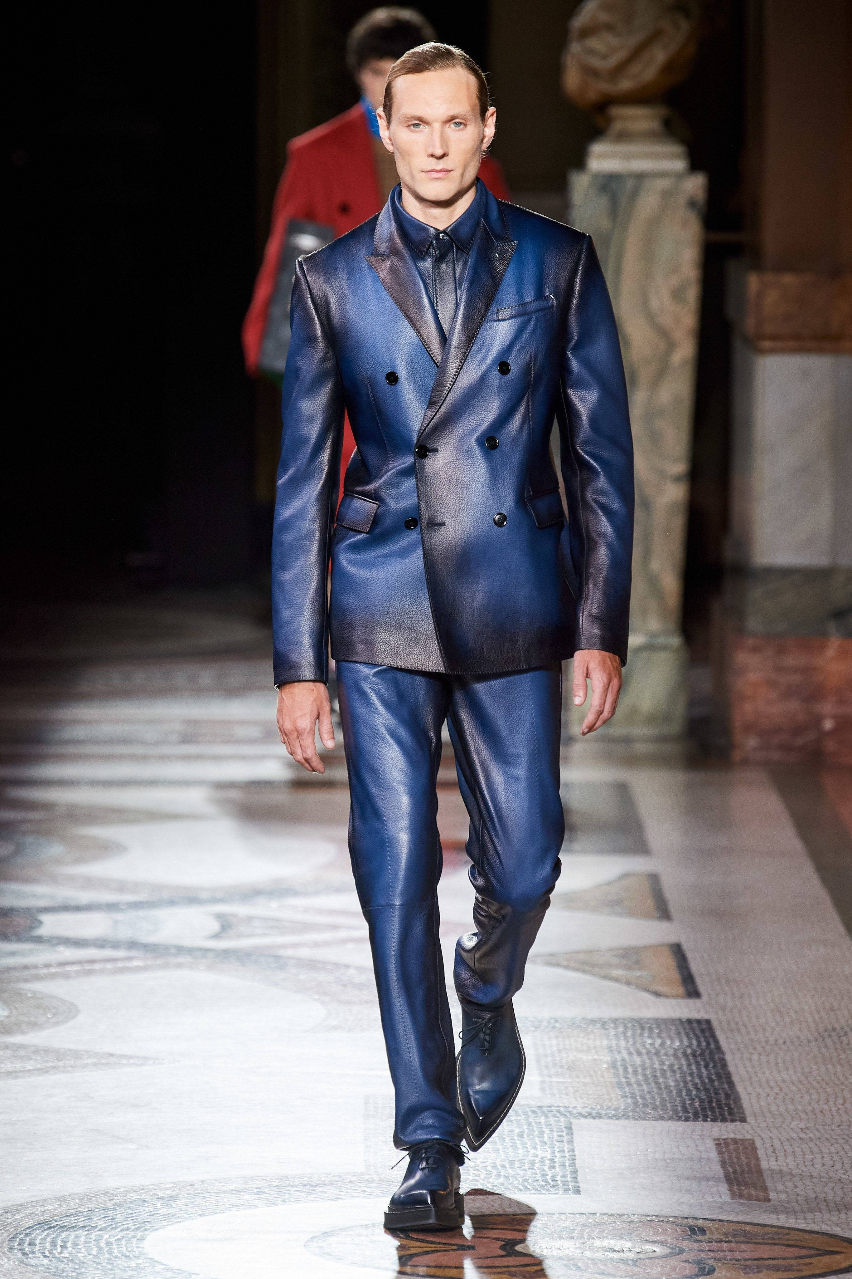Berluti 2020 秋季系列 壁紙 — 一位男模特兒在戲劇性的時裝秀燈光下，自信地穿著閃亮的皇家藍色皮革雙排扣西裝走在雲石天橋上。