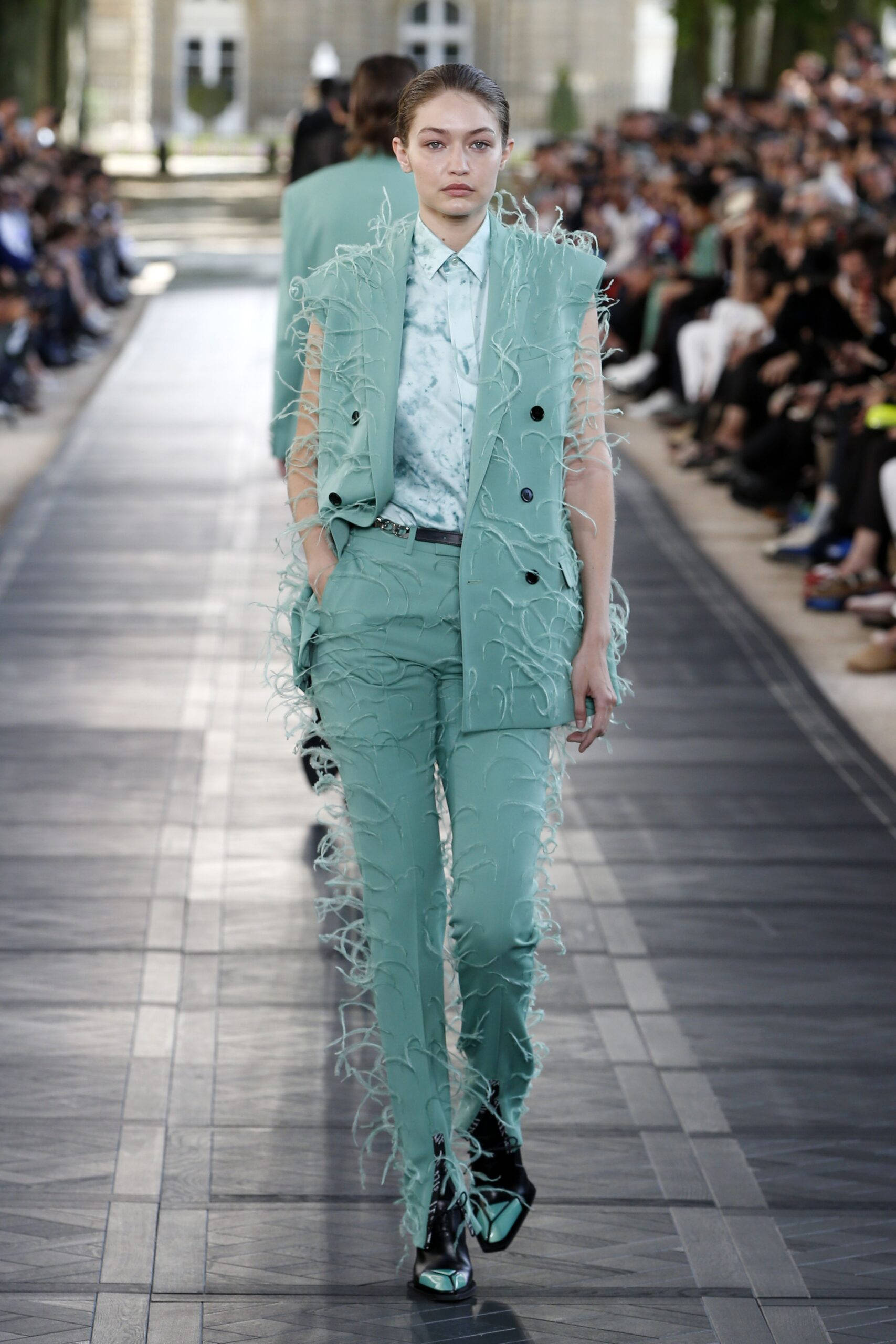 Berluti 2020夏季系列 壁紙 — 一位模特兒穿著帶有羽毛狀流蘇的紋理薄荷綠色西裝和黑色靴子走在天橋上。