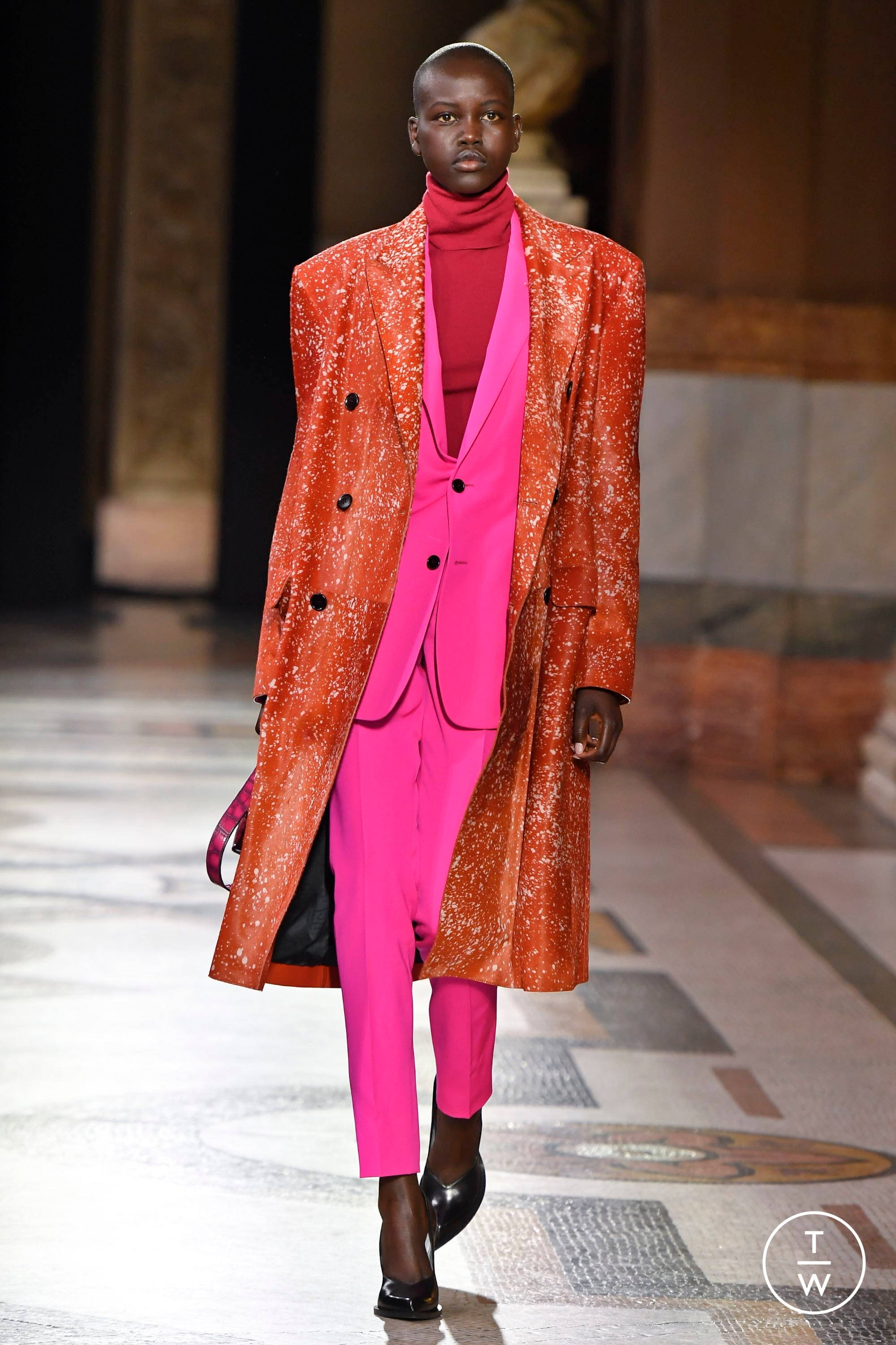 Berluti 橙色外套 壁紙 — 一位模特兒自信地走在天橋上，身穿大膽的橙色閃亮外套，內搭鮮豔的粉紅色西裝和高領毛衣。
