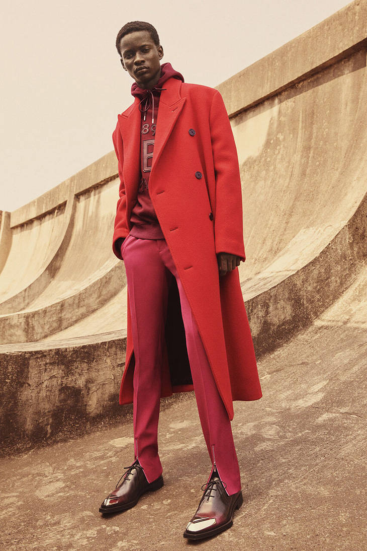 Berluti 紅色外套 壁紙 — 一位時尚男士穿著大膽的紅色外套和配套褲子，在柔和日光下自信地站在彎曲的混凝土牆前。