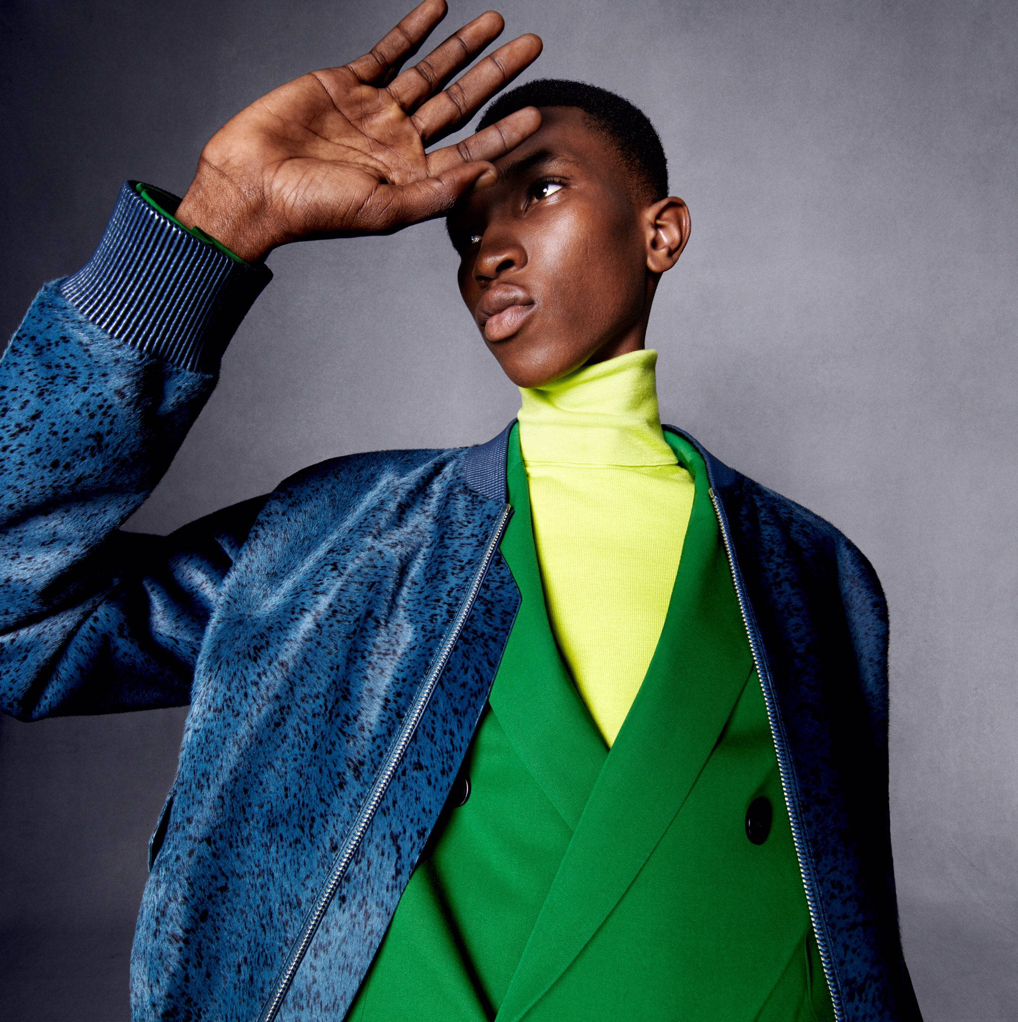 Berluti 冬季 2020 系列 壁紙 — 一位時尚的年輕男子穿著藍色斑點外套和螢光綠色高領毛衣，在灰色攝影棚背景下用手遮擋眼睛擺姿勢。