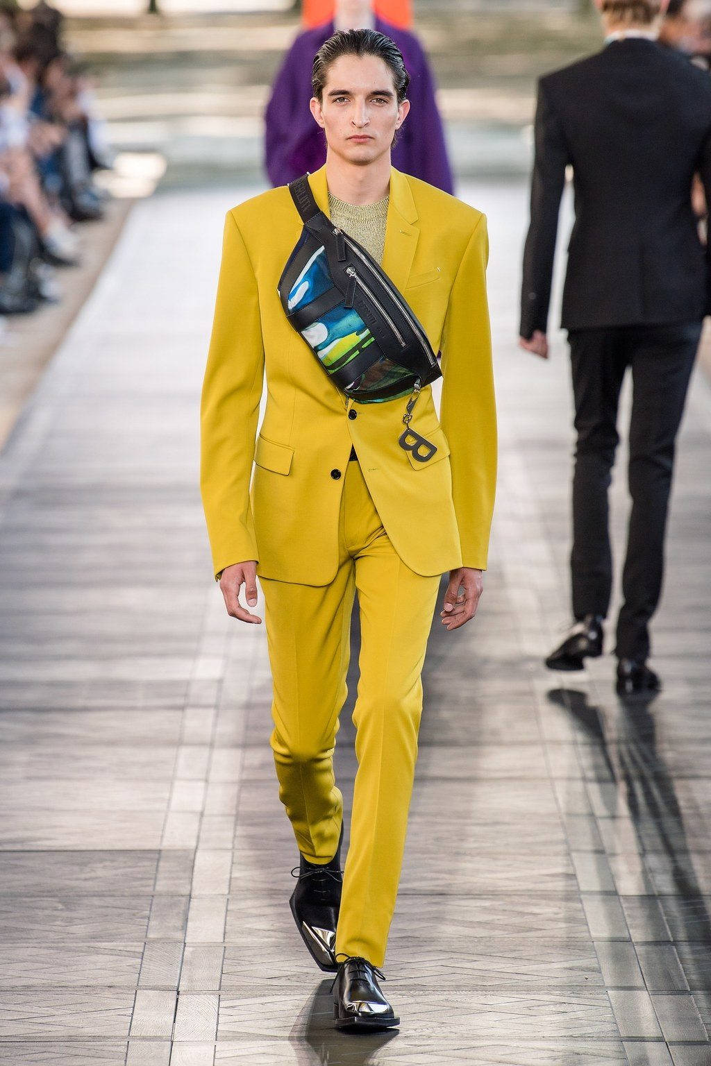Berluti 淺黃色西裝 壁紙 — 一位模特兒穿著大膽的黃色西裝，搭配色彩繽紛的圖案腰包，走在天橋上。