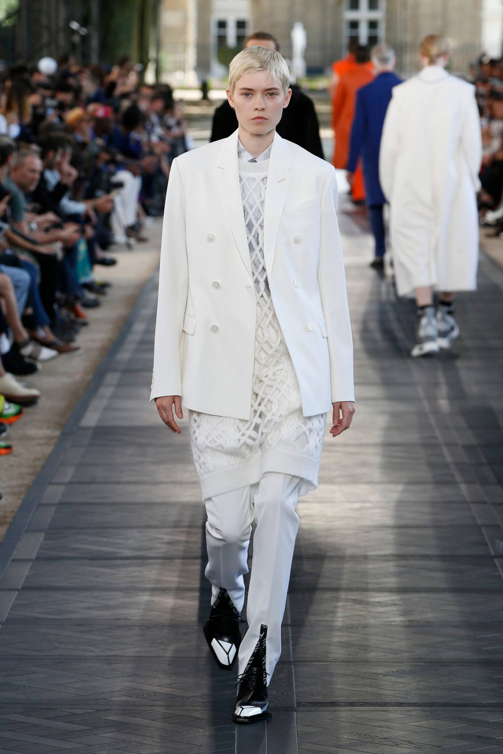 Berluti 白色西裝 壁紙 — 一位短金髮模特兒自信地走在天橋上，身穿白色雙排扣西裝外套和配套長褲，搭配黑白靴子。