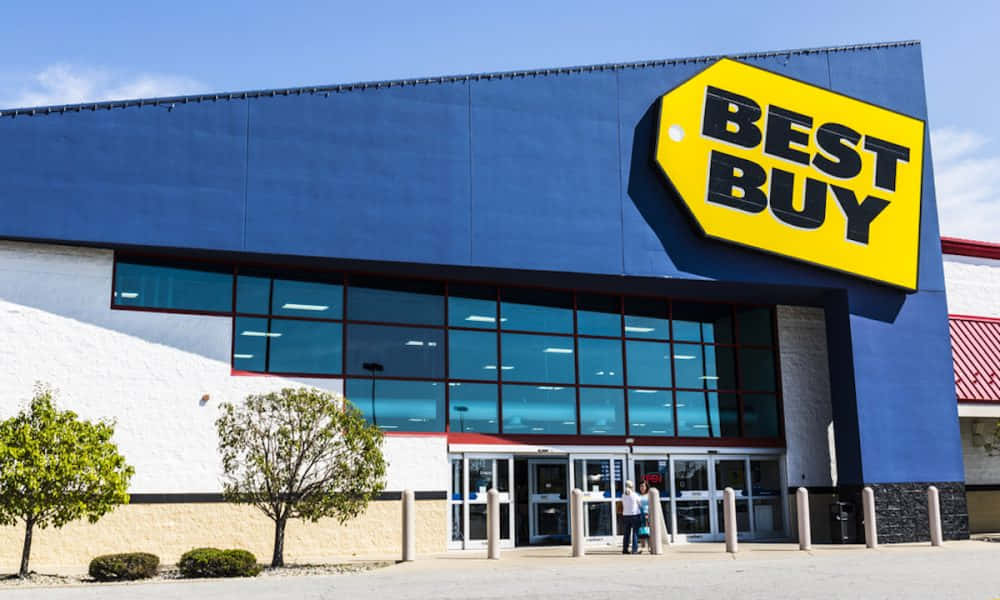為您的家配備所需科技產品——Best Buy 壁紙 — 一間明亮的藍色Best Buy商店，帶有大型黃色標籤標誌，在晴朗的天空下，有兩個人正在進入。