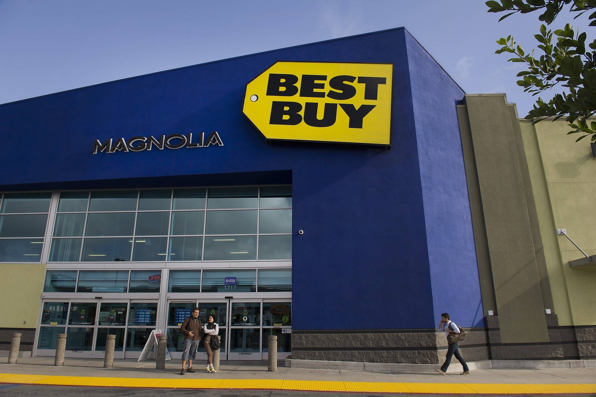 Best Buy 馬格諾利亞店 壁紙 — 晴朗日子，一間亮藍色Best Buy商店，黃色招牌和玻璃門，人們走過。