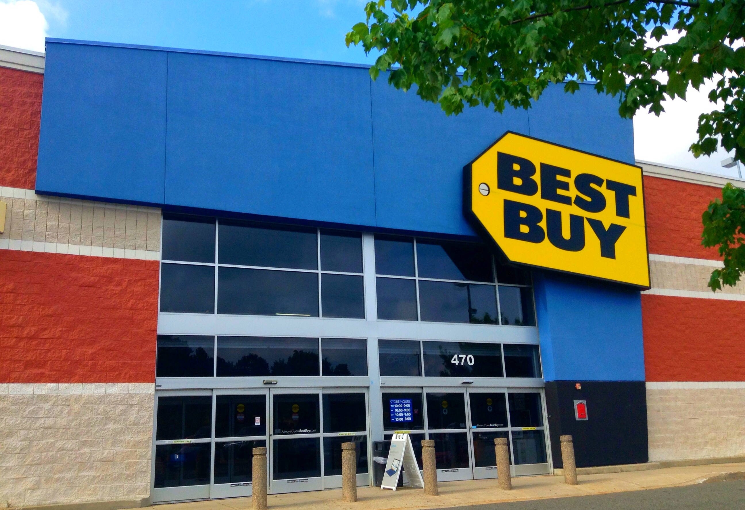 Best Buy乾淨外觀 壁紙 — 一間明亮的藍色和紅色Best Buy商店，帶有大型黃色標誌，在晴朗的天空下，樹枝框住右上角。