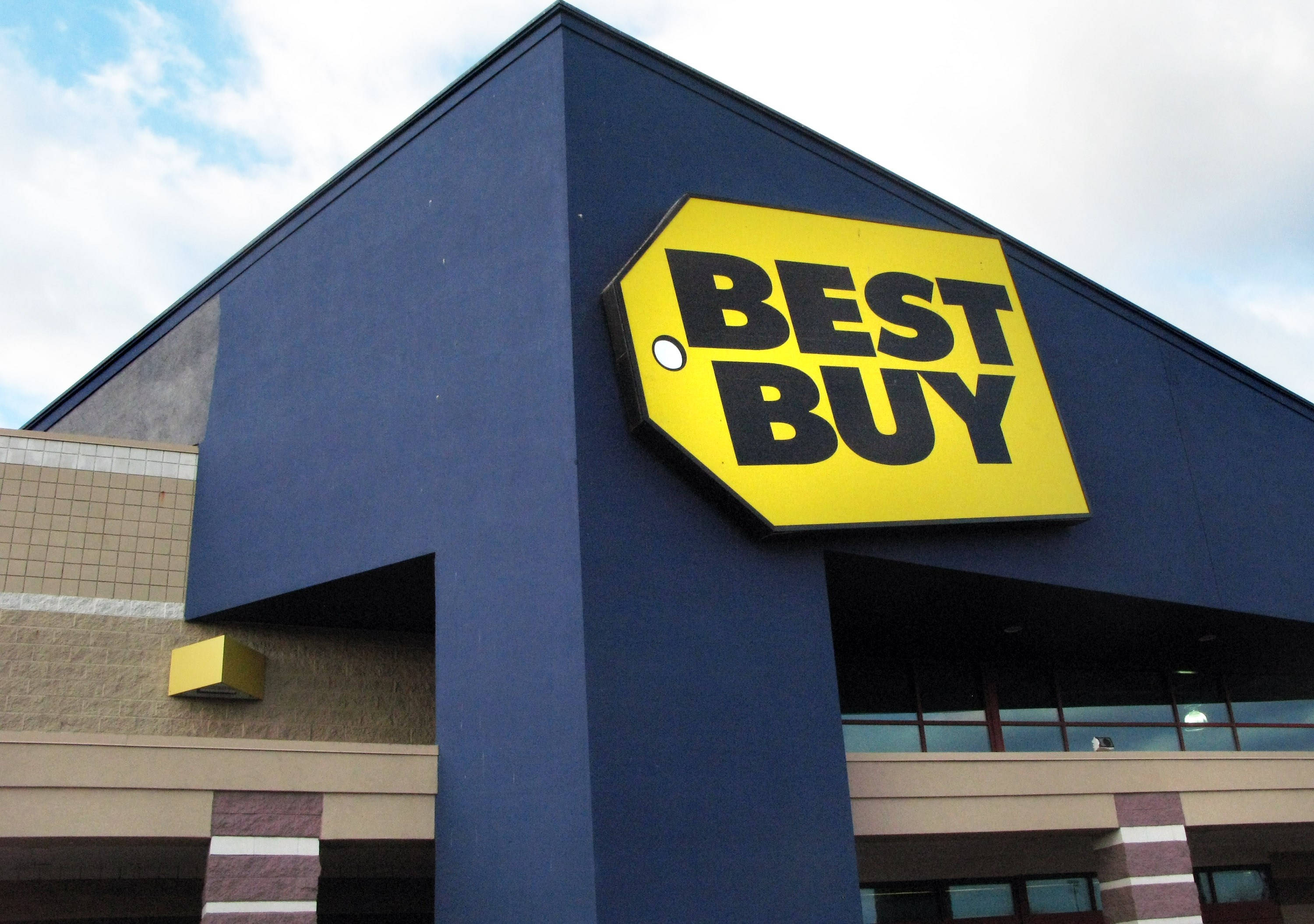 Best Buy建築招牌 壁紙 — 在部分多雲的天空下，一個大膽的黃色Best Buy標誌，帶有黑色字體，安裝在深藍色建築物外牆上。