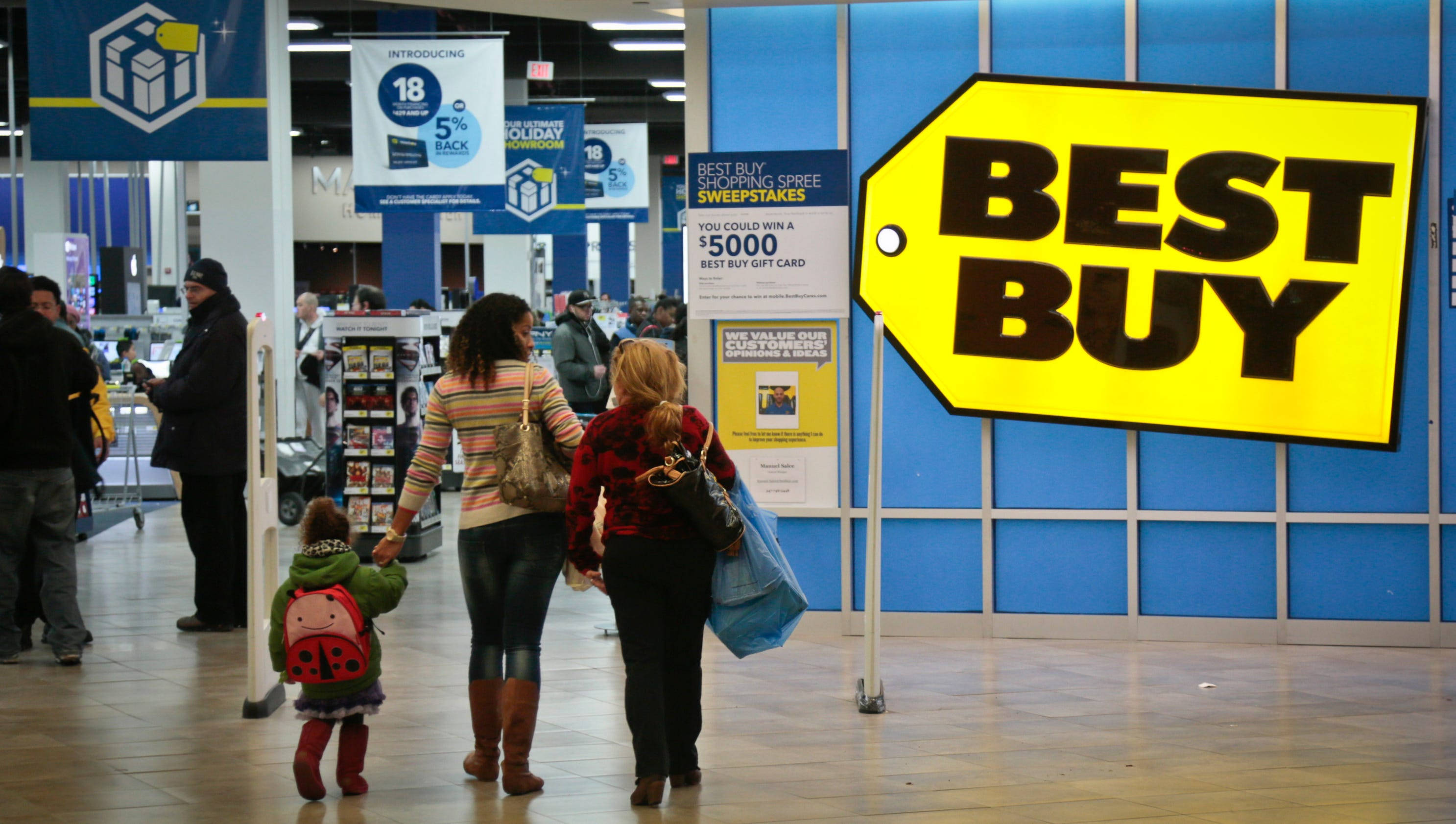 Best Buy購物中心 壁紙 — 購物者走過光線明亮的 Best Buy 商店，頭頂上有大型黃色標籤標誌和促銷橫幅。