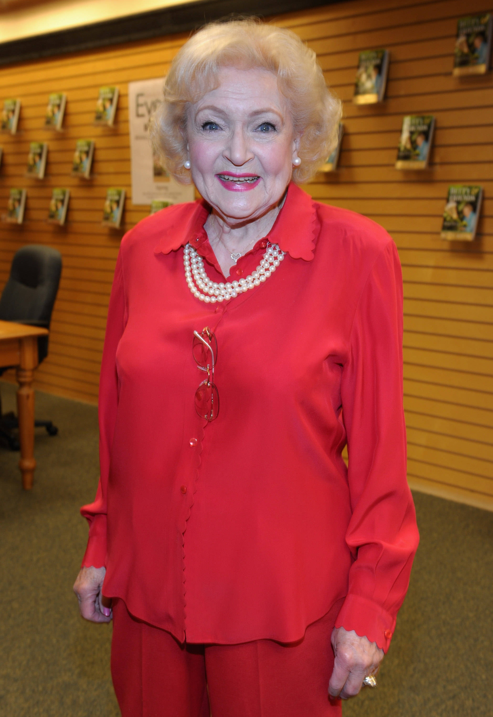 Betty White 簽書會 壁紙 — 伊芙·阿登穿著鮮紅色西裝和珍珠項鍊，在木書架背景下溫暖地微笑。