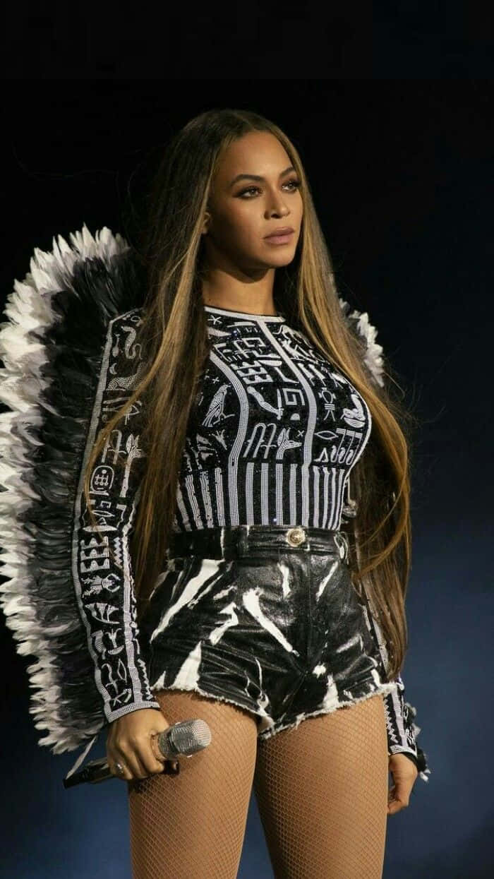 Beyoncé 桌布 壁紙 — Beyoncé 自信地站在舞台上，身穿帶有象形文字圖案的黑白羽毛天使服裝，手持麥克風。