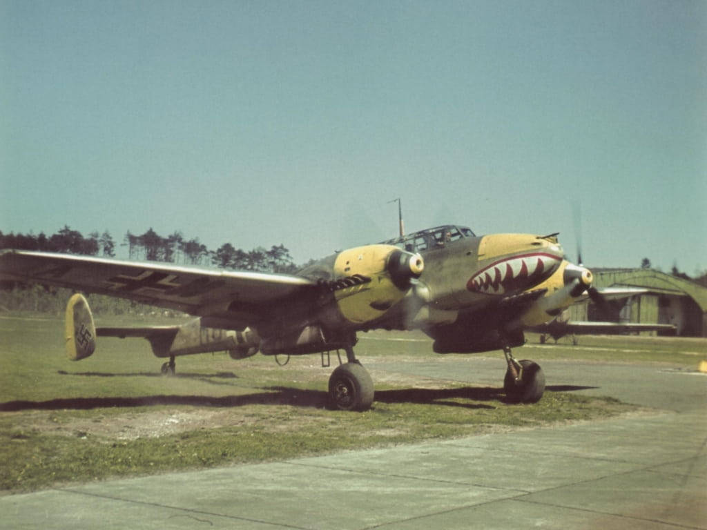 Bf 110德國二戰戰鬥機舊照 壁紙 — 復古軍用飛機帶有鯊魚嘴鼻藝術，停在停機坪上，藍天白雲，靠近機庫。