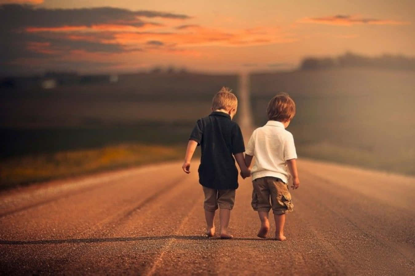 BFFs：最佳朋友永遠相伴 壁紙 — 兩個赤腳的小孩手牽手在日落時分沿著鄉村道路行走，天空呈現溫暖的橙色和粉紅色。