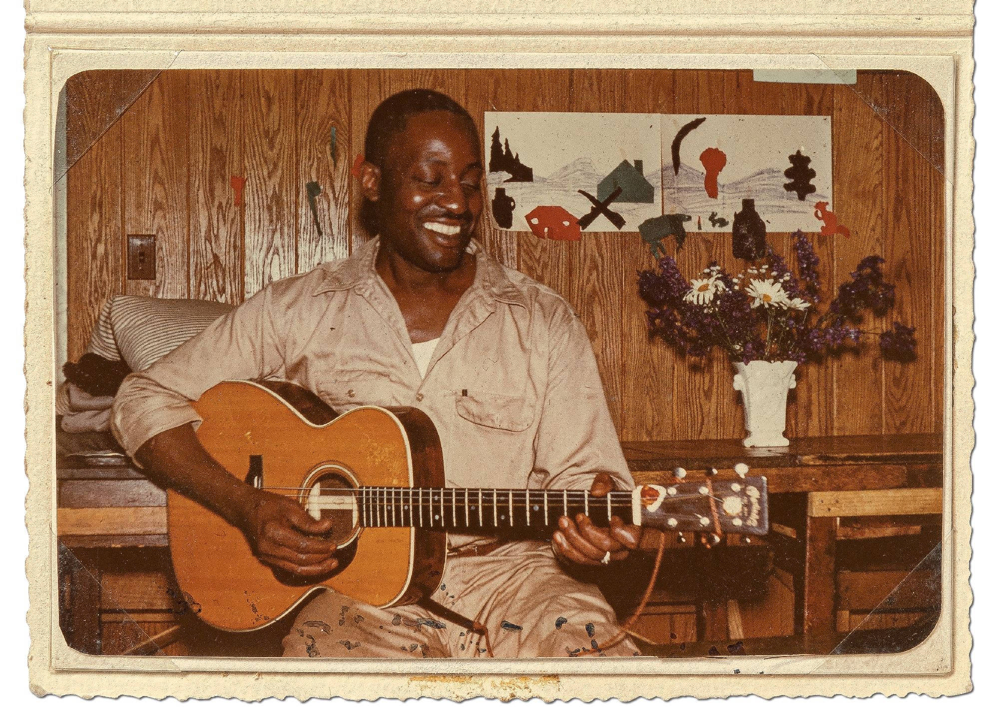 美國藝術家 Big Bill Broonzy 壁紙 — 一個快樂的男人在舒適的木鑲板房間裡，一邊彈奏木結他，一邊微笑，旁邊放著一瓶花。