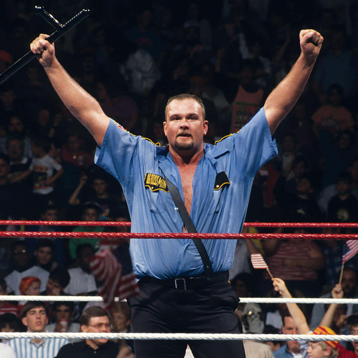 Big Boss Man 雙臂高舉 壁紙 — 一位肌肉發達的摔角手身穿藍色制服，在燈光明亮的擂台上，在歡呼的人群面前勝利地舉起拳頭和警棍。