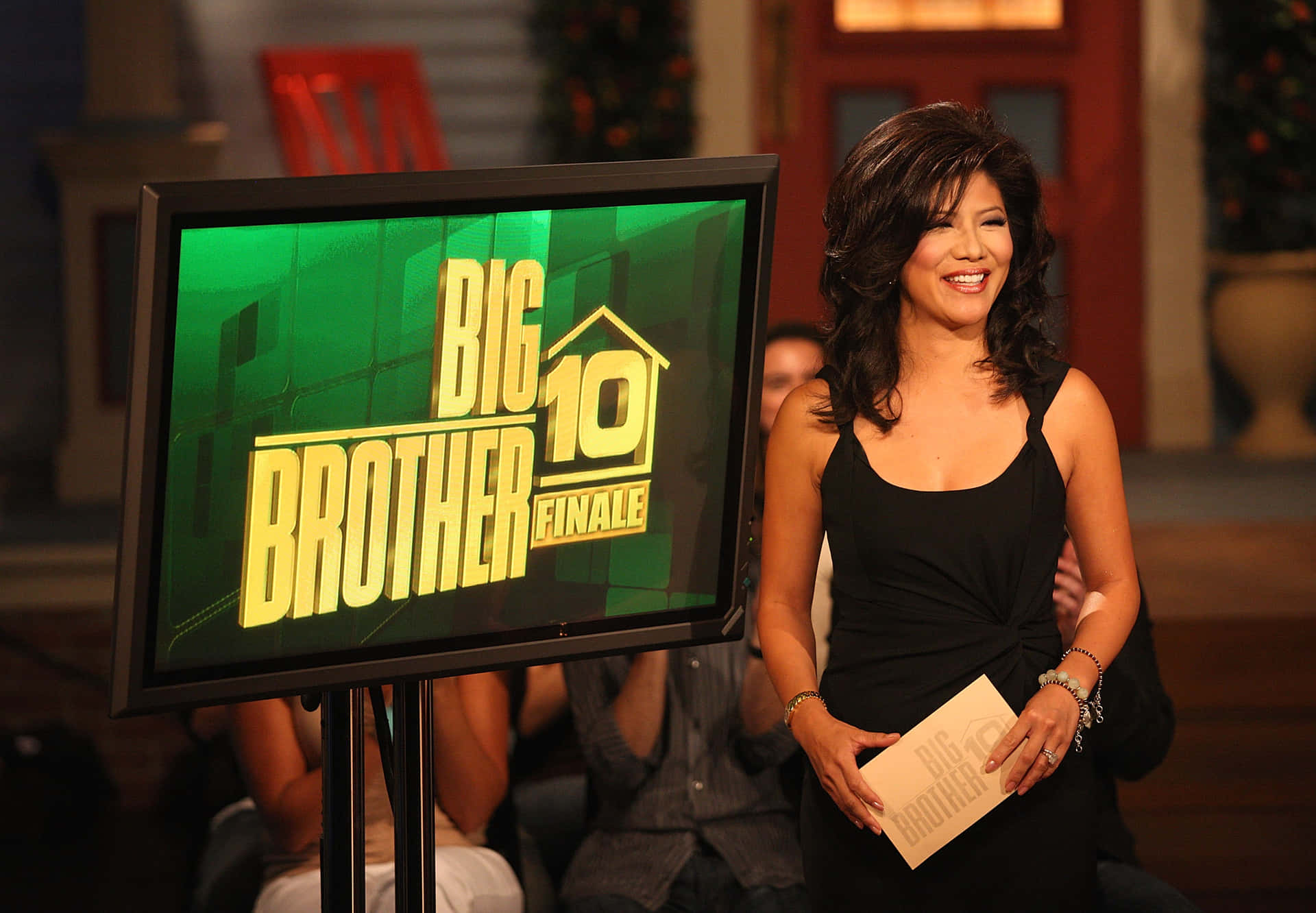 壁紙 — 一位微笑的黑裙女士站在顯示「Big Brother 10 Finale」的屏幕旁，文字為綠色背景上的粗體黃色。