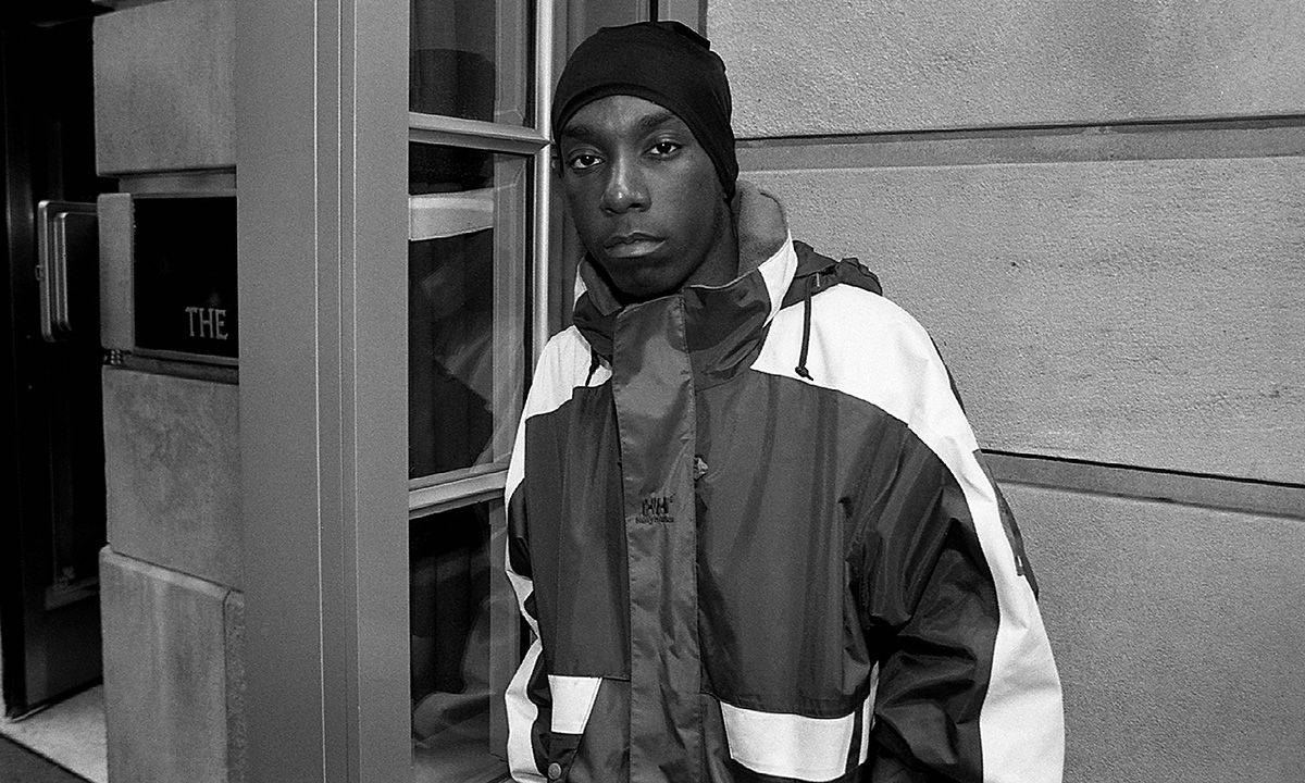 Big L 黑白影像 壁紙 — 一位年輕男子穿著大膽的雙色外套和冷帽，自信地站在單色調的混凝土建築外牆前。