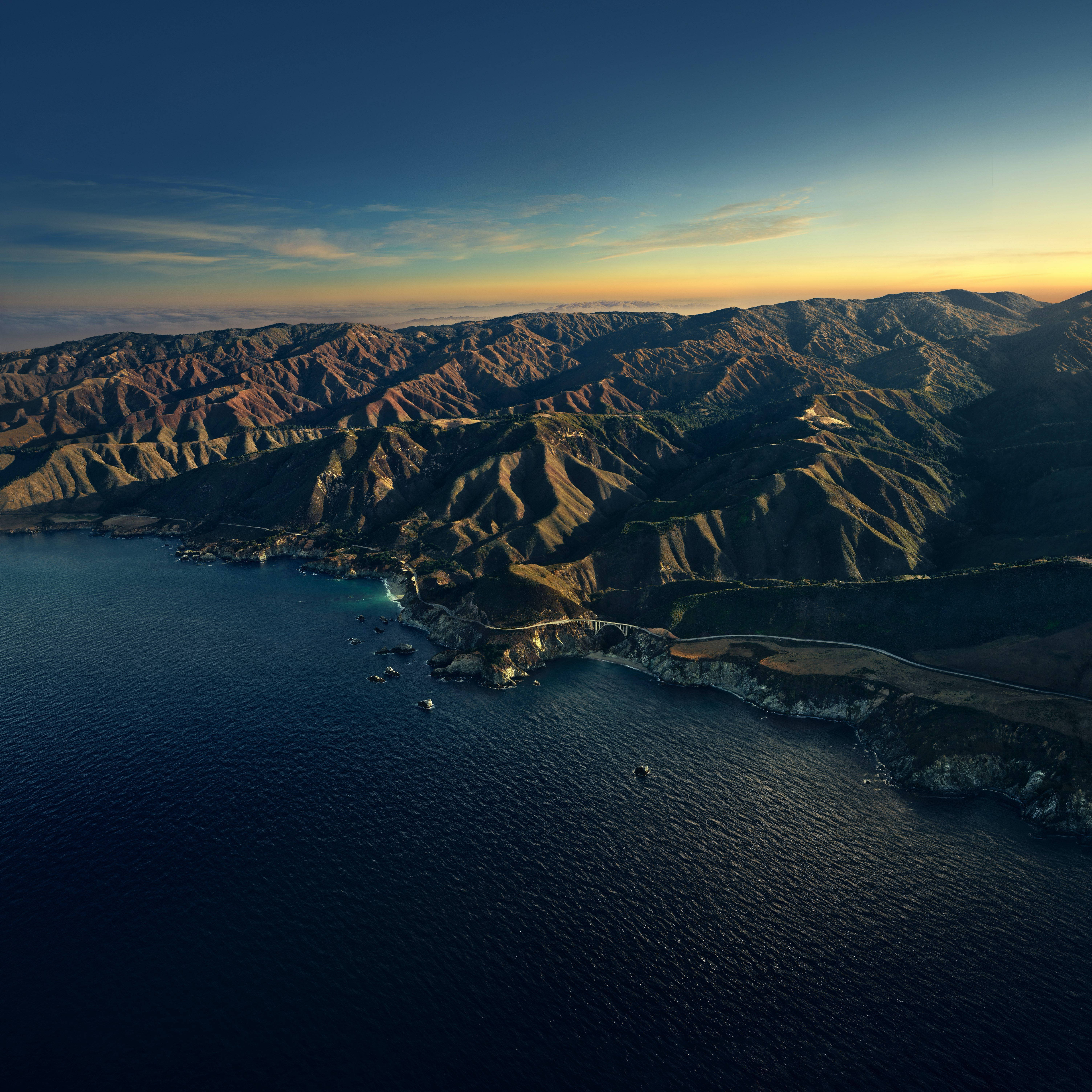 Big Sur 山巒日落地平線 壁紙 — 崎嶇海岸山脈的鳥瞰圖，沐浴在金色日落光芒中，下方是深藍色的海洋。