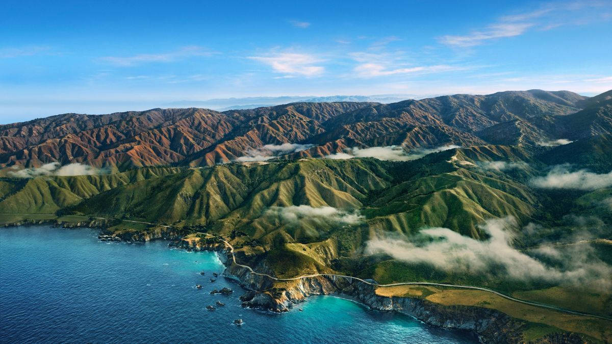 Big Sur 綠山與低垂雲霧 壁紙 — 崎嶇的綠色山脈與碧綠海洋交匯的鳥瞰圖，蜿蜒的海岸道路和清澈藍天下飄浮的雲朵。