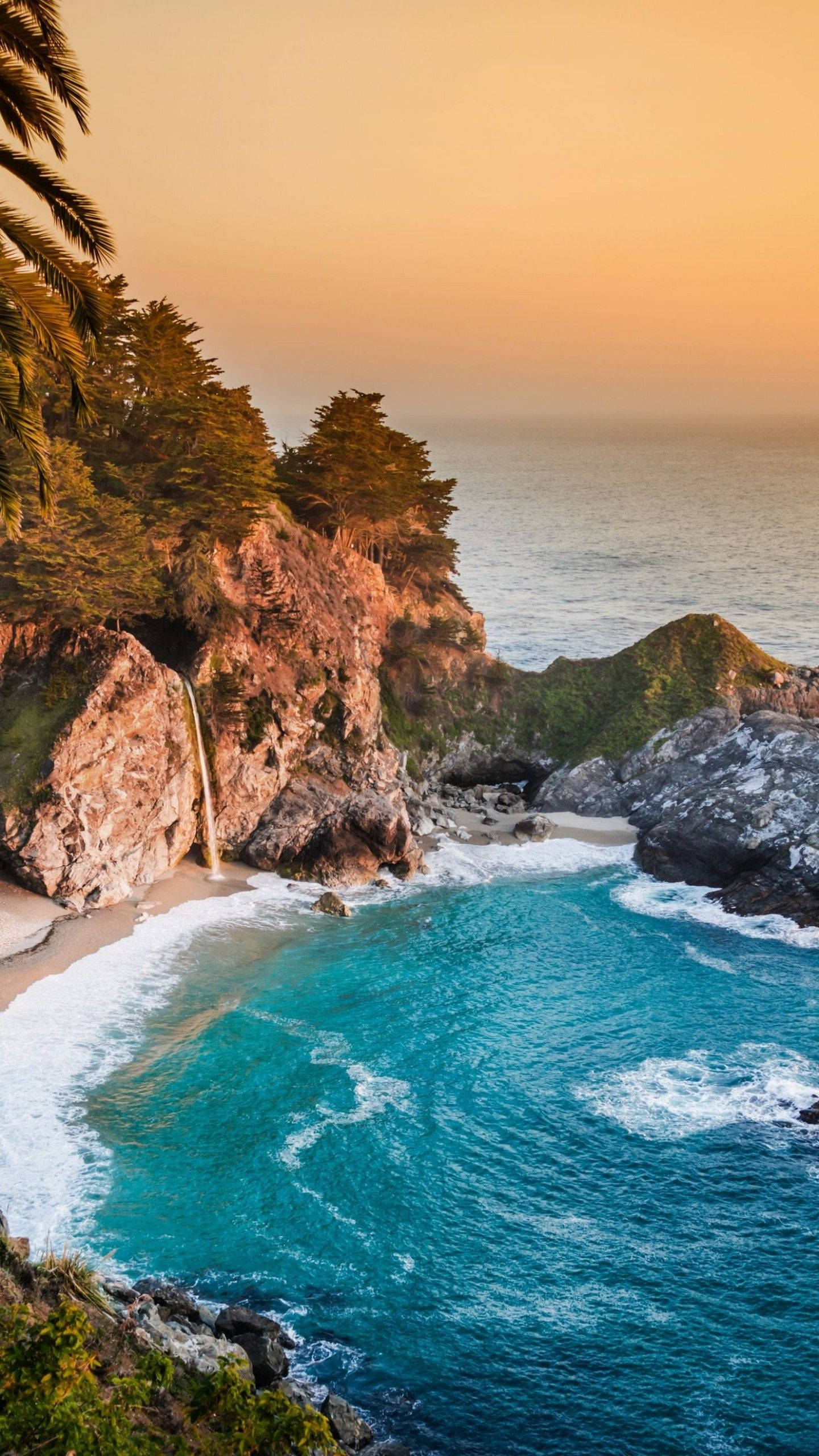 Big Sur 藍色潟湖手機壁紙 壁紙 — 日落時分，一個令人驚嘆的海岸小灣，碧綠的海水拍打著岩石海岸，瀑布從棕櫚樹環繞的懸崖上傾瀉而下。