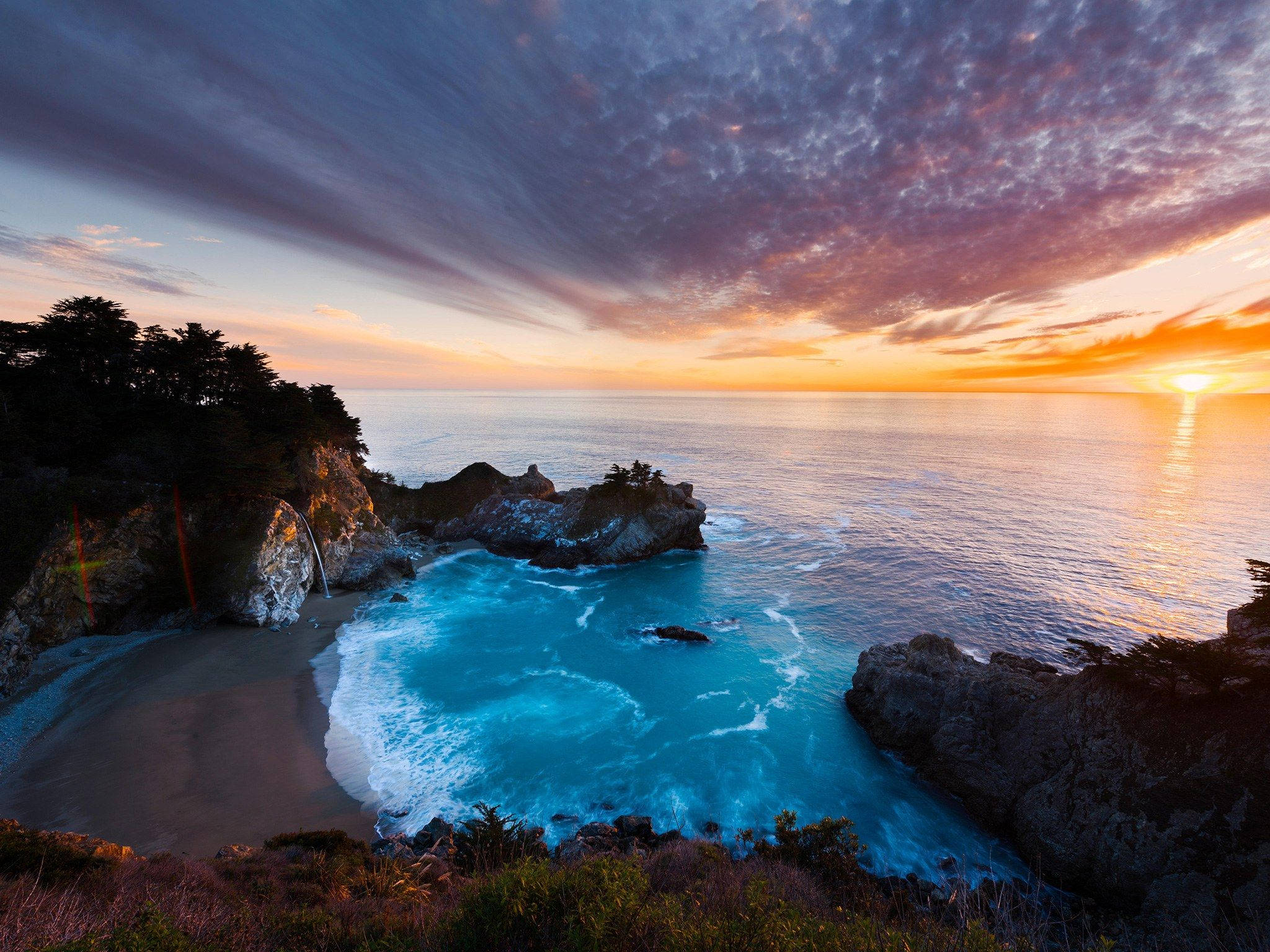 Big Sur 雲霞與日落 壁紙 — 一個令人嘆為觀止的日落，位於一個僻靜的海灣，碧綠的海水拍打著崎嶇的懸崖，一個小瀑布傾瀉到沙灘上。