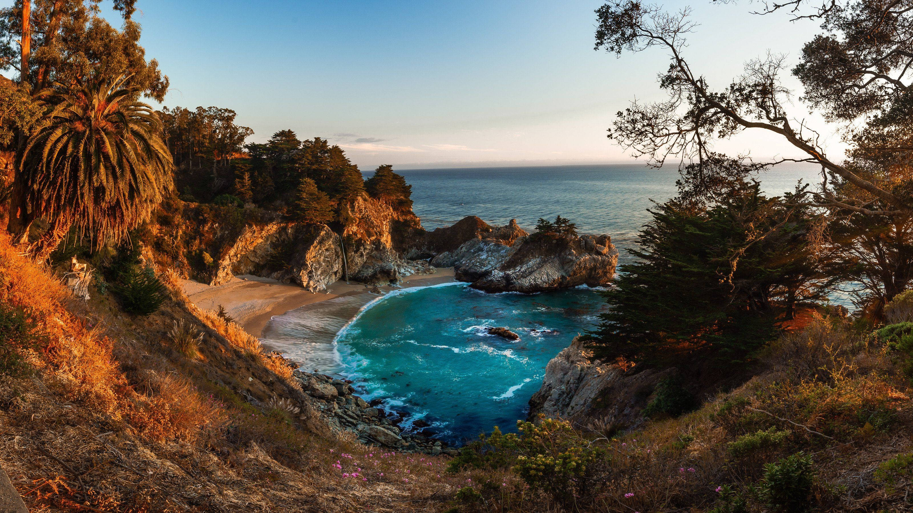 Big Sur 麥威瀑布與潟湖 壁紙 — 金色夕陽灑落在一個僻靜的海灣，碧綠的海水、崎嶇的懸崖，四周被茂密的樹木環繞。