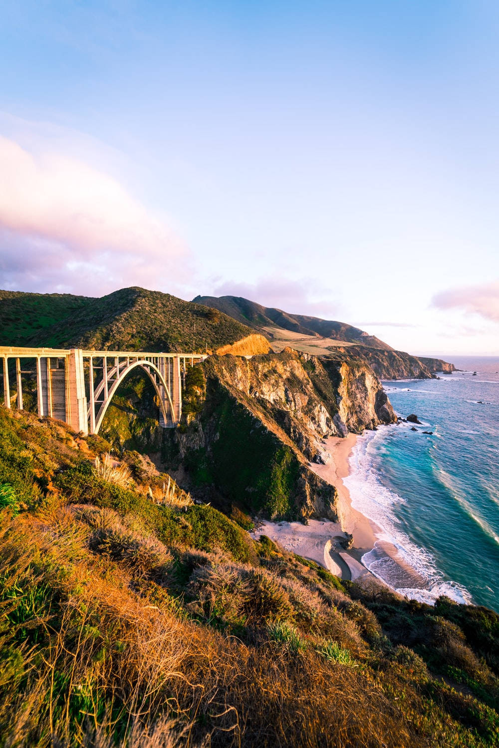 Big Sur 黃金時刻自然風光 壁紙 — 日落時分，一座雄偉的拱橋橫跨崎嶇的海岸懸崖，下方碧綠的海浪拍打著沙灘。