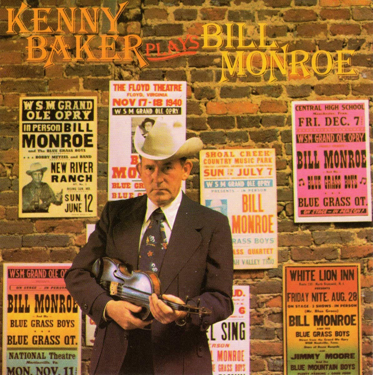 Bill Monroe 紀念：Kenny Baker 演奏小提琴 壁紙 — 一名身穿西裝和牛仔帽的男子，拿著小提琴靠在一面貼滿復古藍草音樂會海報的磚牆上。