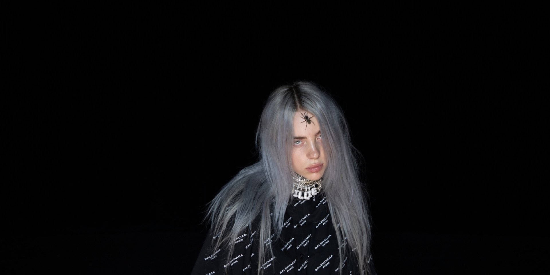 Billie Eilish 穿著 Balenciaga 衫 壁紙 — 一個長著銀色長髮、額頭上有蜘蛛紋身的人，在黑色背景下強烈地凝視著。