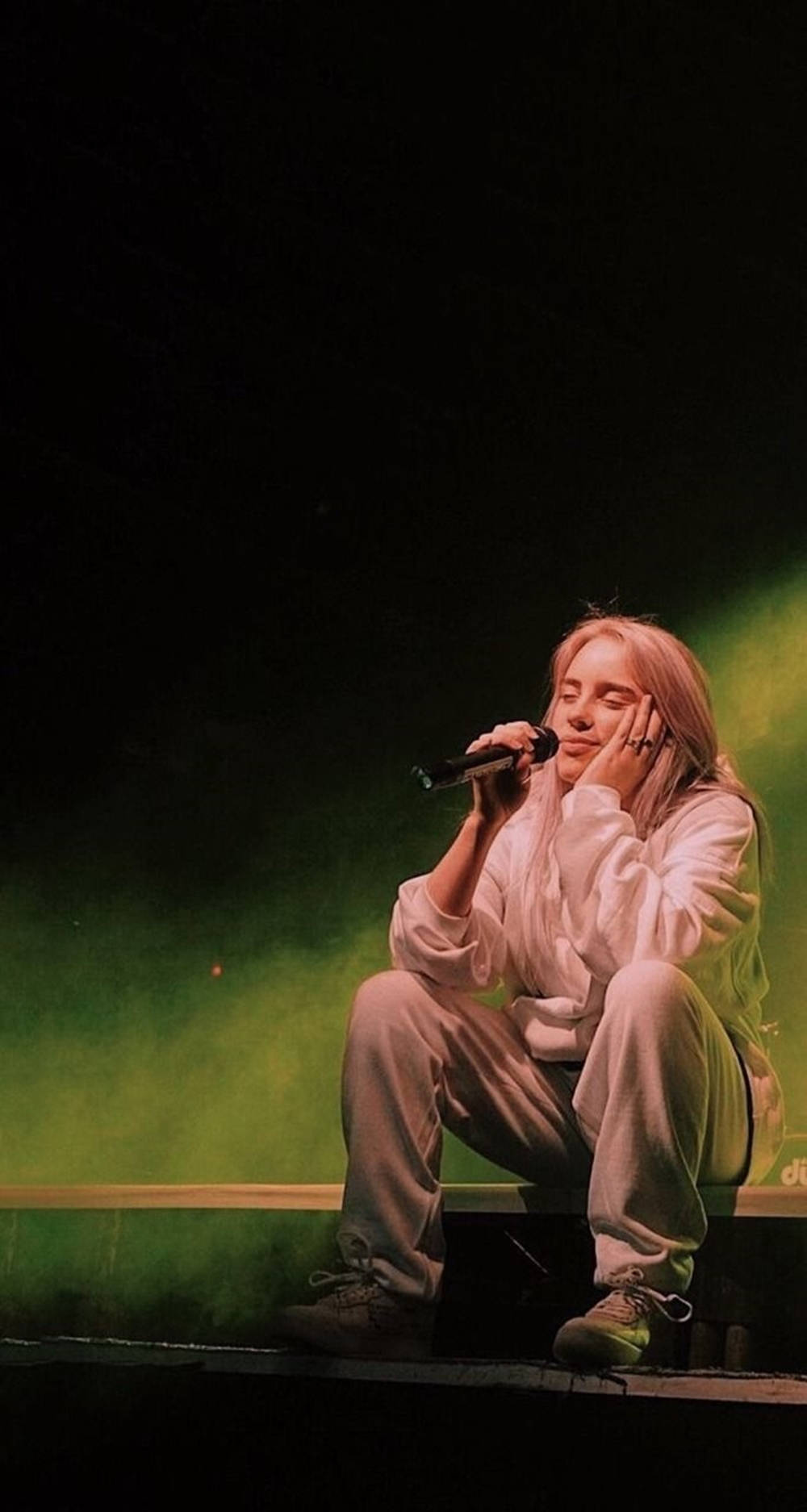 Billie Eilish 美學舞台坐姿壁紙 壁紙 — 一位金髮表演者坐在舞台上，手持麥克風，被綠色舞台燈光照射，背景黑暗。