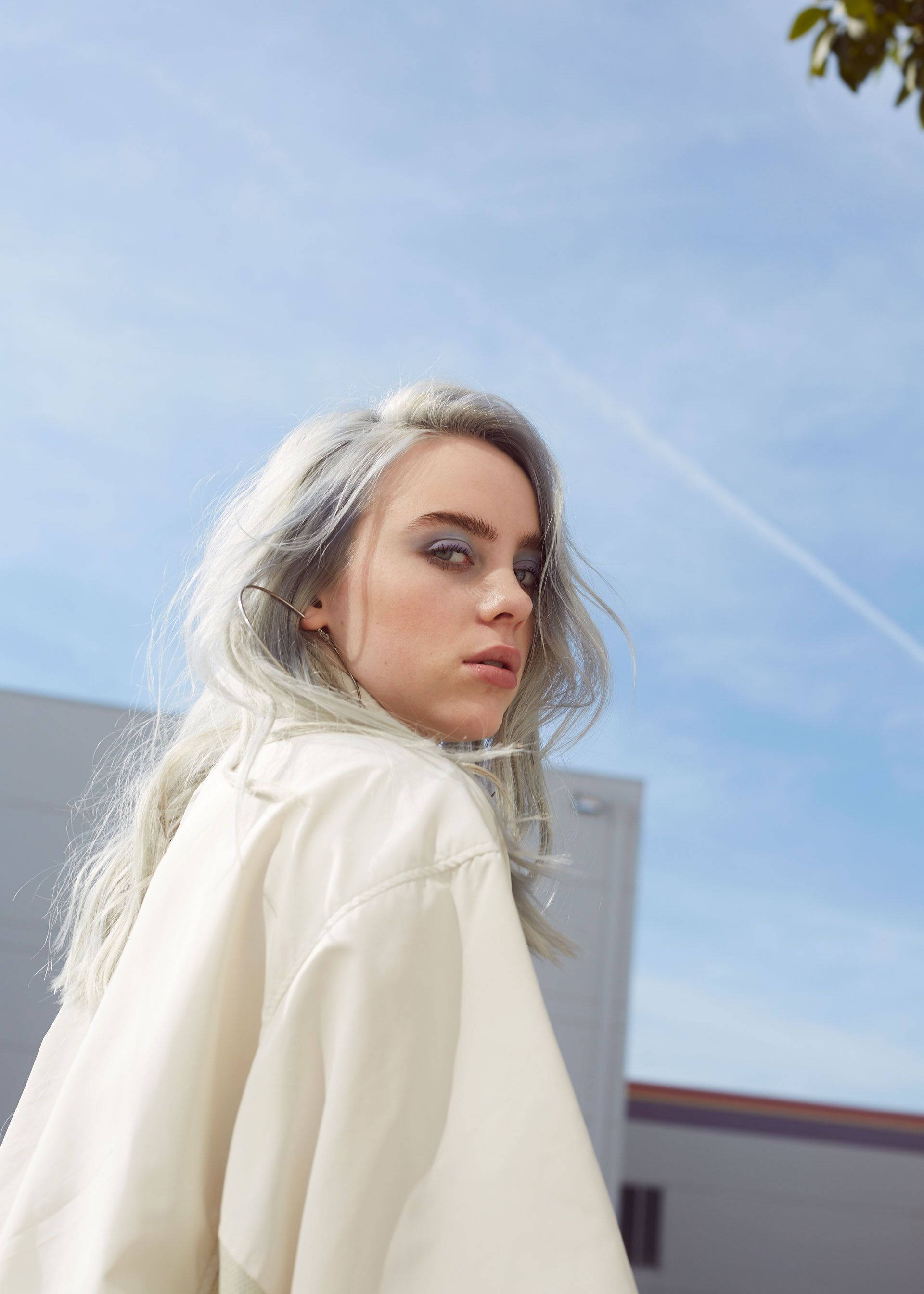 Billie Eilish 黃色襯衫天空背景美學照 壁紙 — 一位年輕女性，銀色頭髮，大膽妝容，在藍天白雲下回頭凝視。