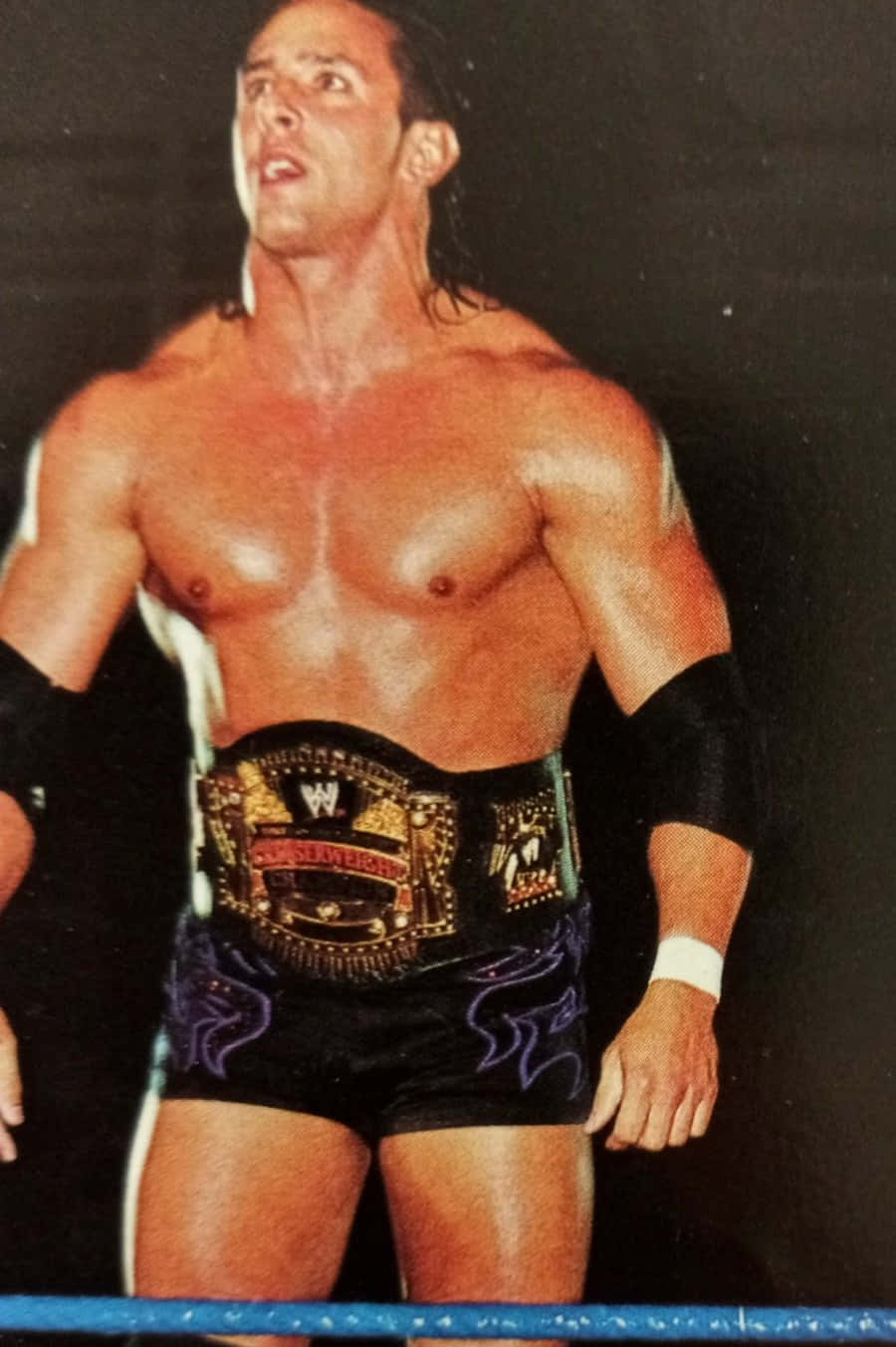 Billy Kidman 身披冠軍腰帶 壁紙 — 一位肌肉發達、長黑髮的摔角手站在擂台上，戴著冠軍腰帶，穿著帶有紫色火焰圖案的黑色短褲。