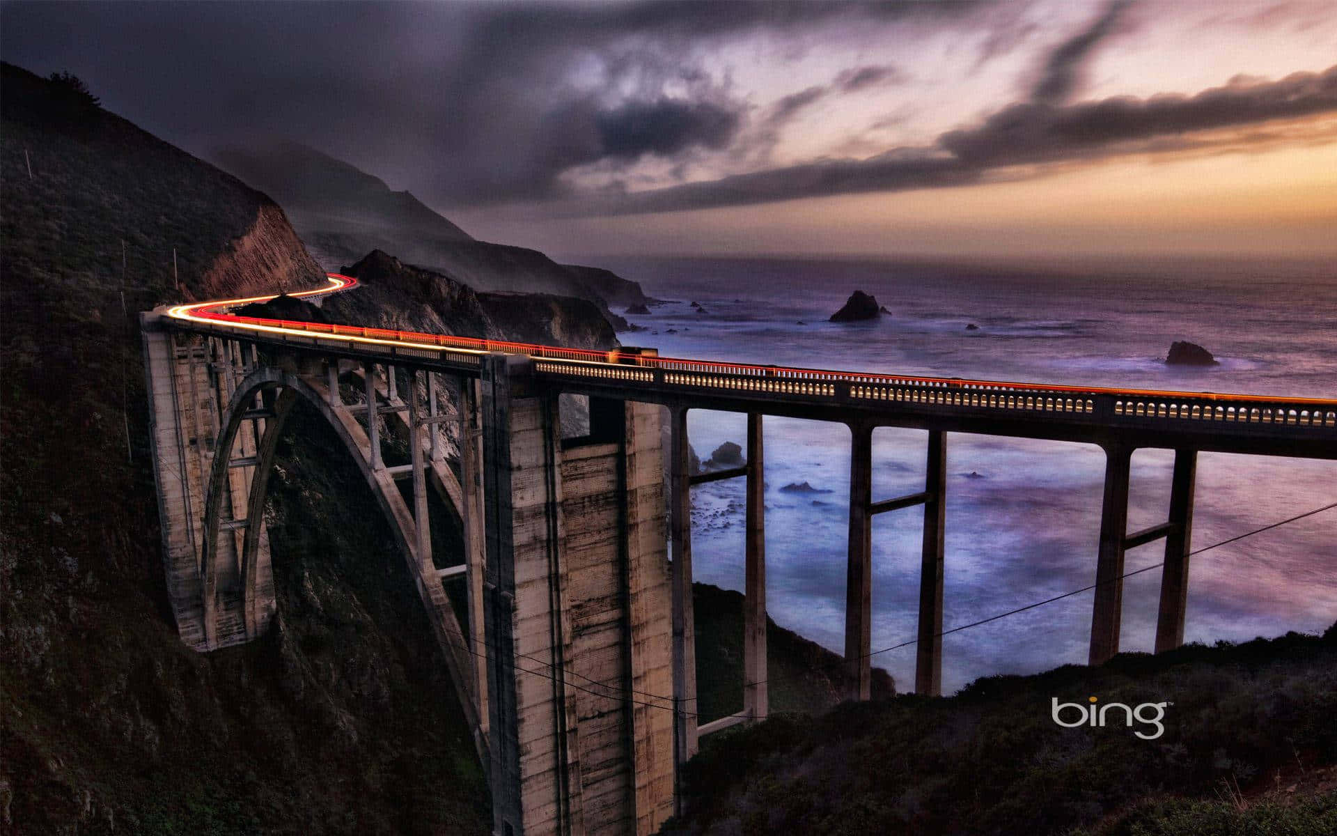Bing 以繽紛美麗的背景領先潮流 壁紙 — 一列發光的火車在暮色中穿越高聳的海岸橋樑，下方波濤洶湧，天空呈現戲劇性的紫色和橙色。