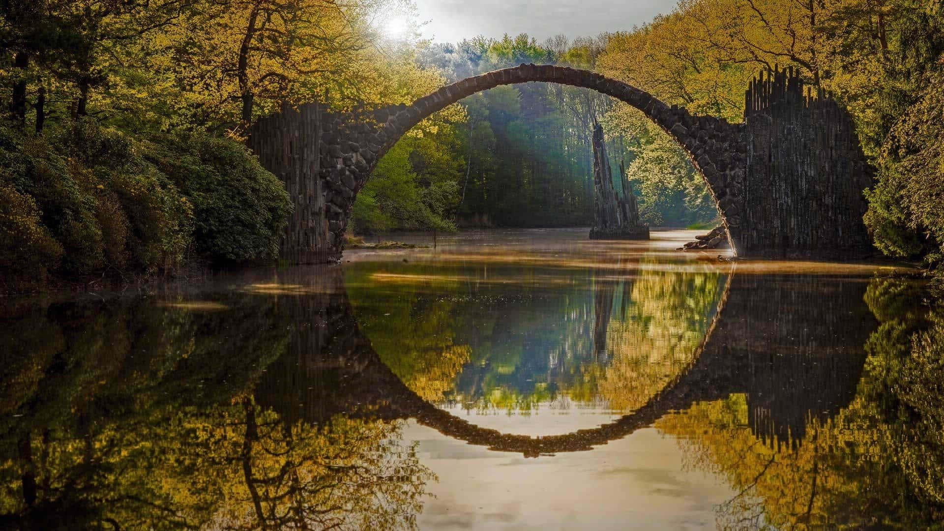 Bing - 發現與探索世界內容 壁紙 — 一座石拱橋橫跨平靜的河流，完美倒映在被秋樹環繞的水面上。