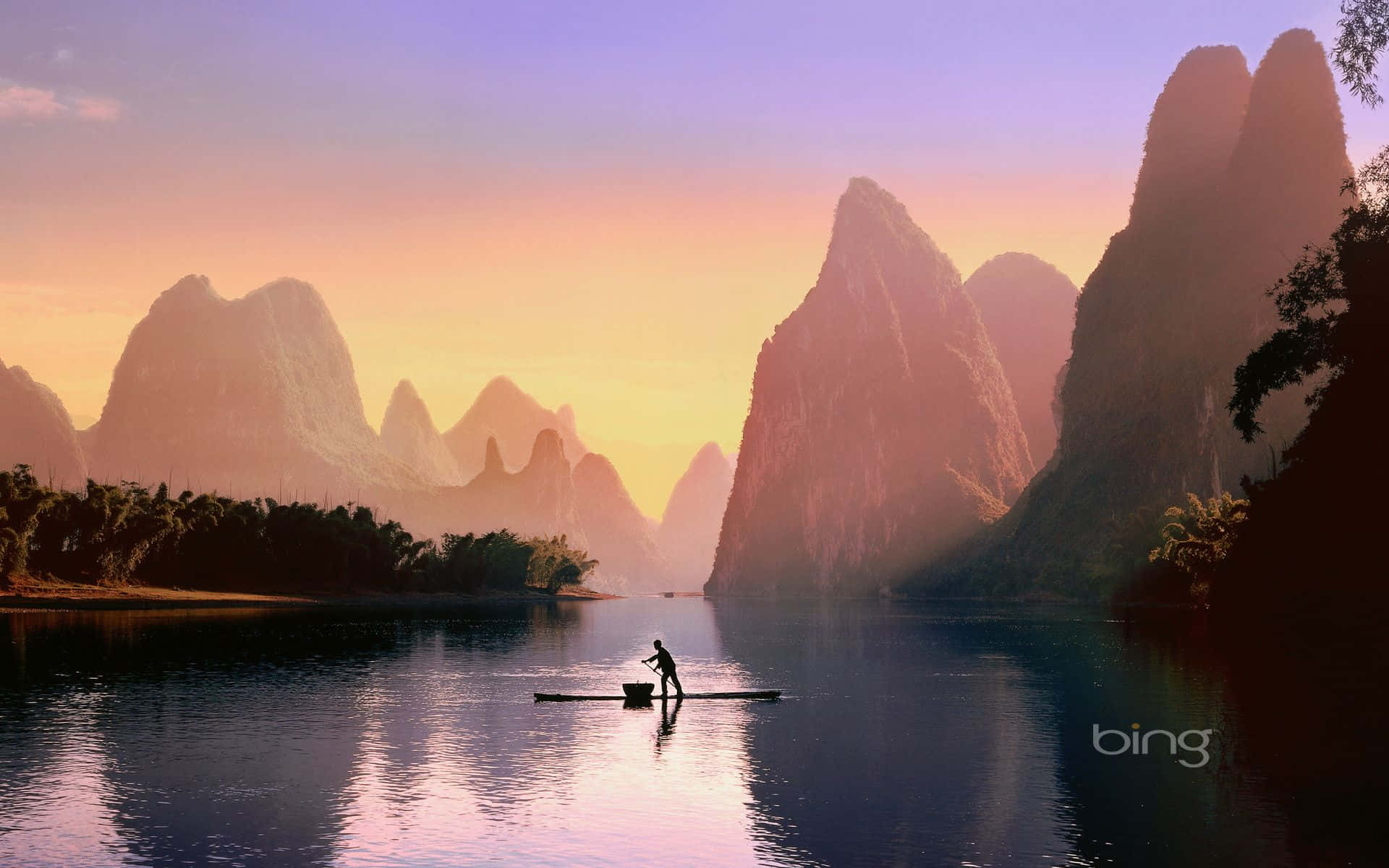 Bing 背景：體驗自然之美 壁紙 — 夕陽西下，一名孤獨的漁夫划著竹筏，穿過寧靜的河流，高聳的喀斯特山峰倒映在平靜的水面上。