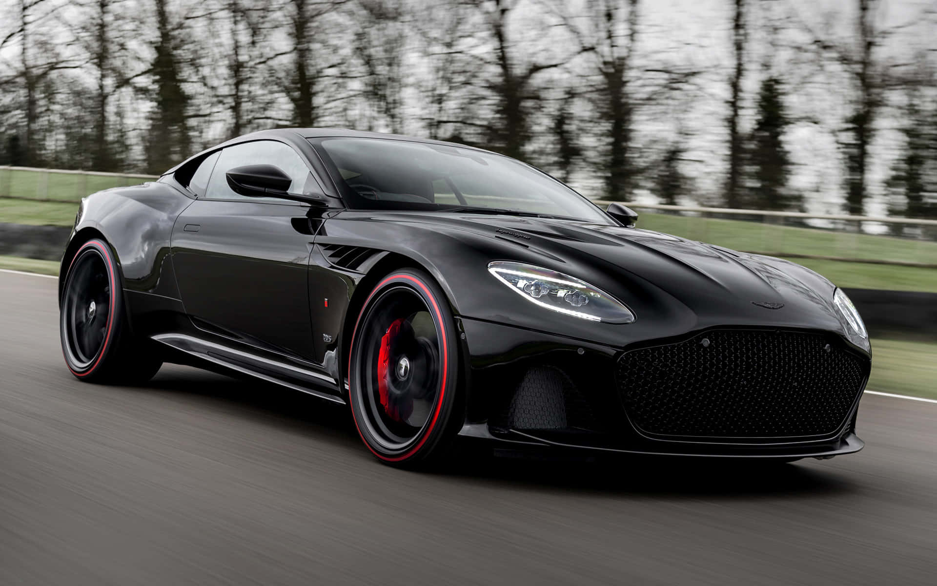 Aston Martin Dbs Superleggera 桌布 壁紙 — 一輛時尚黑色Aston Martin跑車，配以紅色點綴的車輪，在兩旁光禿樹木的賽道上疾馳。