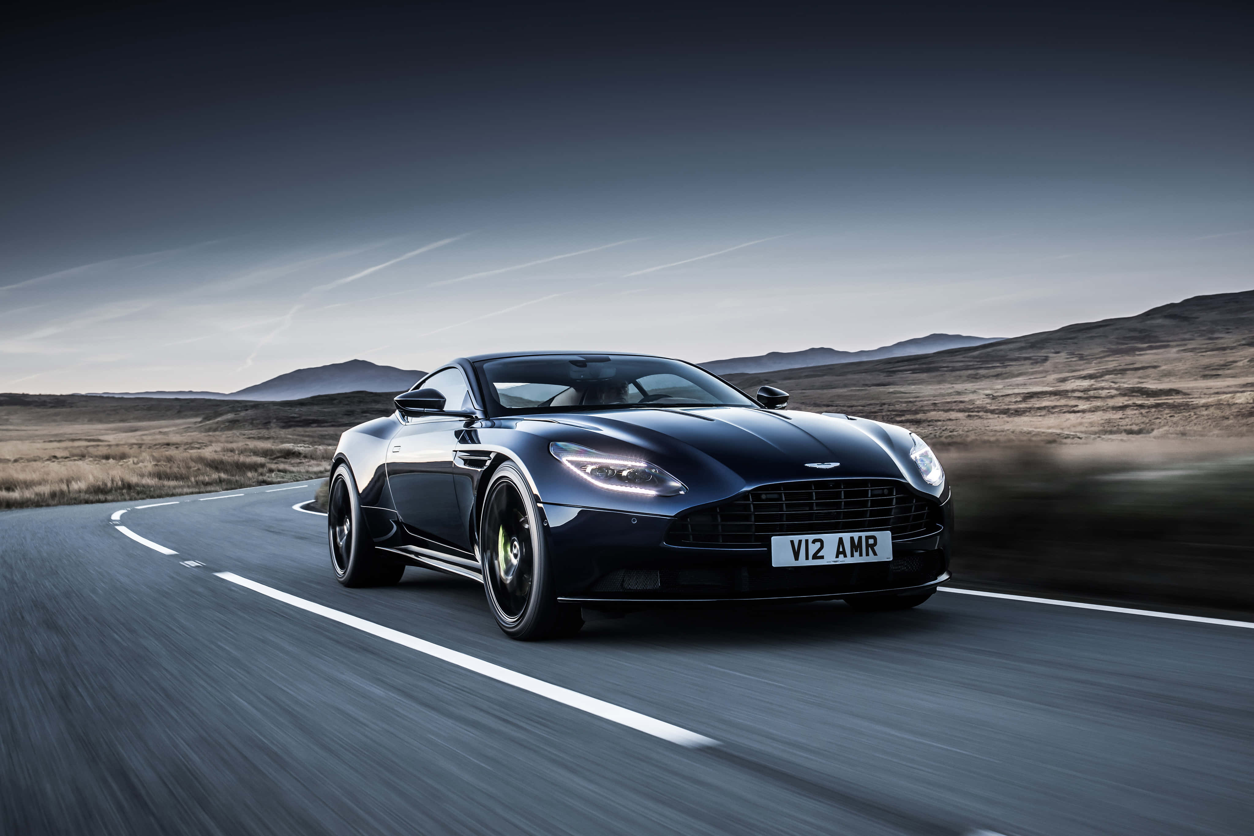 Aston Martin Db11 桌布 壁紙 — 一架流線型的黑色 Aston Martin V12 AMR 在荒涼的山區景觀中沿著蜿蜒的道路奔馳，天空籠罩著陰鬱的暮色。