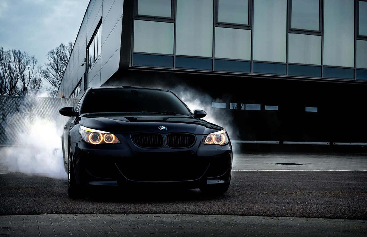 Bmw M5 桌布 壁紙 — 一輛黑色寶馬跑車在陰天下一棟現代玻璃建築前漂移，輪胎冒出滾滾濃煙。