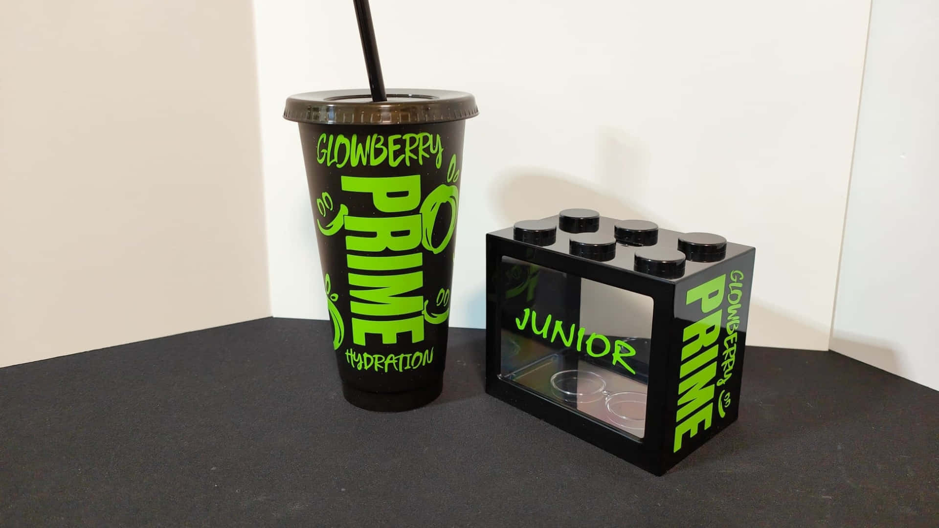  壁紙 — 一個黑色杯子和一個帶有亮綠色 'Glowberry Prime' 品牌的 LEGO 風格盒子，放在深色表面上，背景是純白色牆壁。