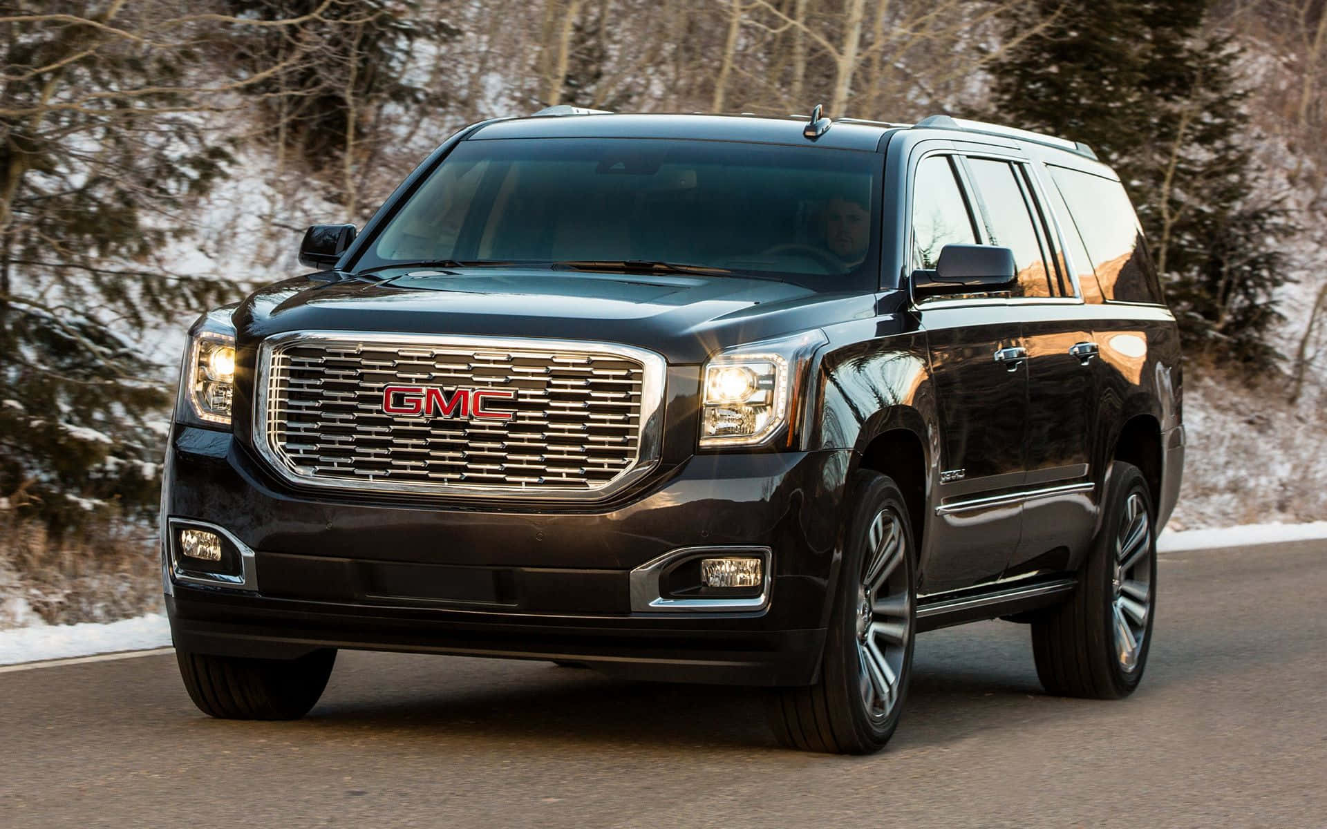 Gmc Yukon 桌布 壁紙 — 一輛黑色GMC Yukon SUV在雪路上行駛，穿過冬季森林，樹木光禿，地面積雪。