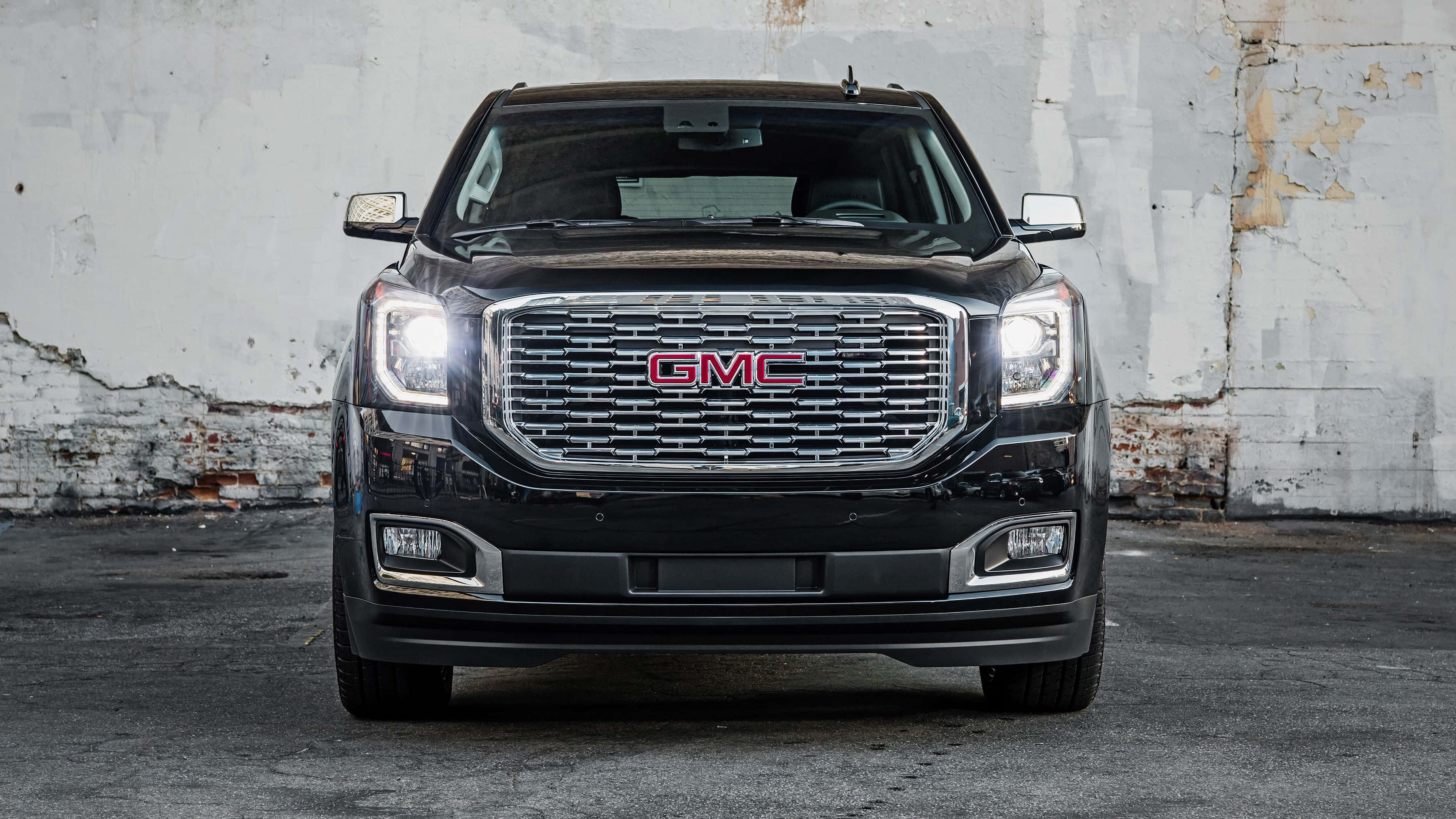 Gmc Yukon 桌布 壁紙 — 一輛黑色GMC育空SUV正面朝向，車頭燈亮起，背景是風化的混凝土牆。