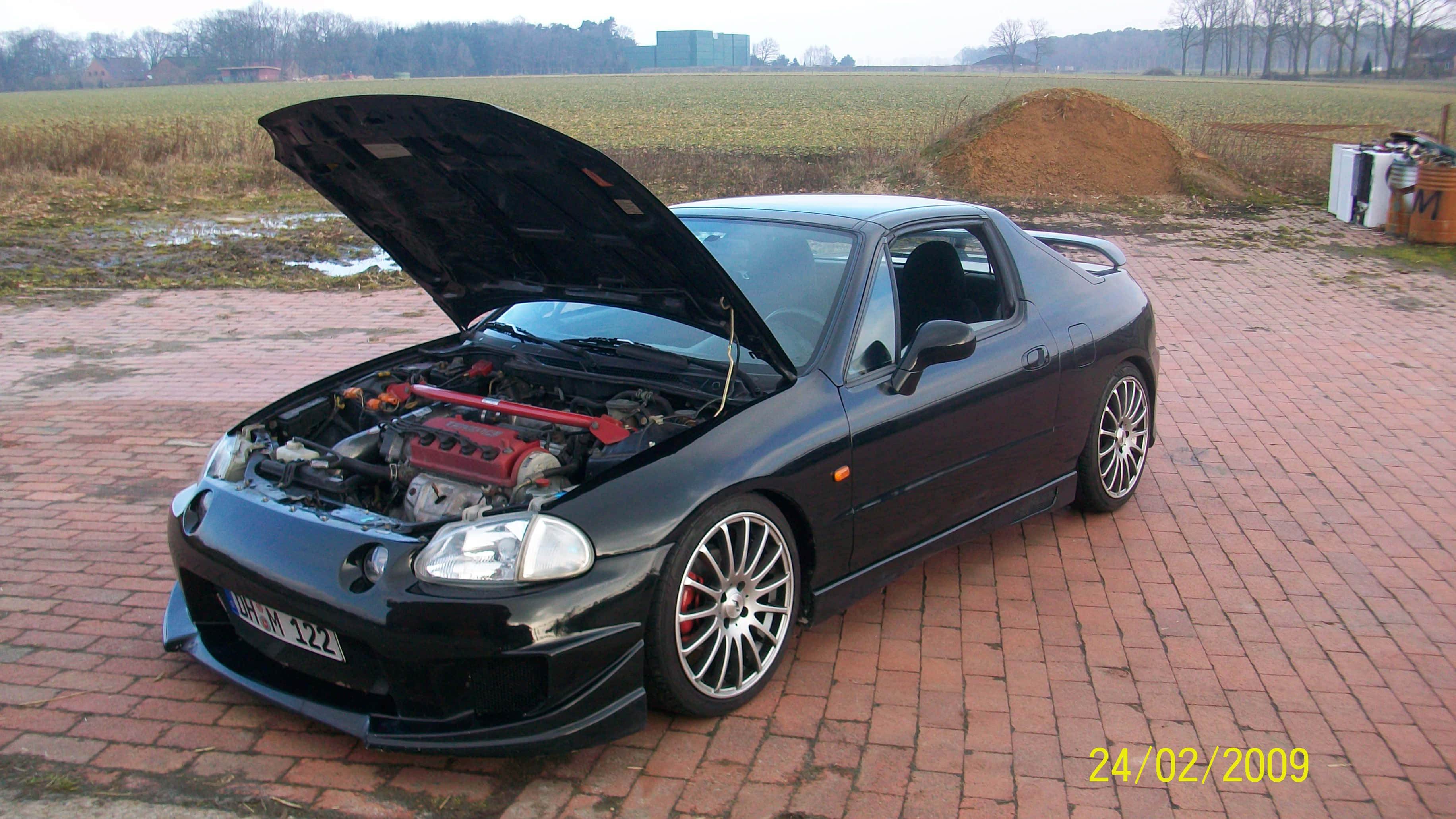 Honda Civic Del Sol 桌布 壁紙 — 一輛黑色跑車打開引擎蓋，露出紅色引擎，停在鄉村田野的磚砌車道上。