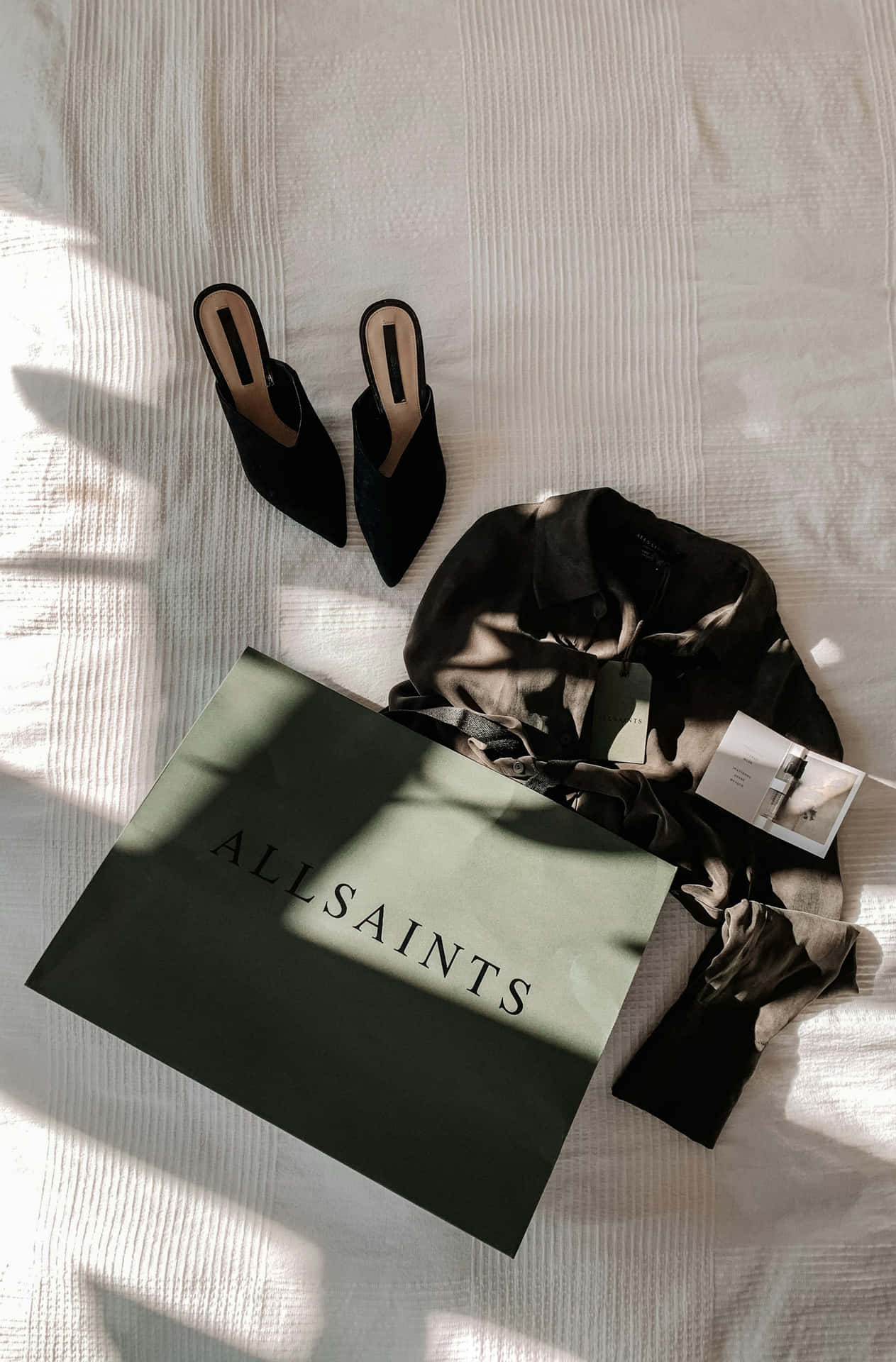  壁紙 — 黑色天鵝絨穆勒鞋和深色襯衫放在AllSaints手袋旁邊，位於陽光照射的白色床罩上，帶有條紋質感。