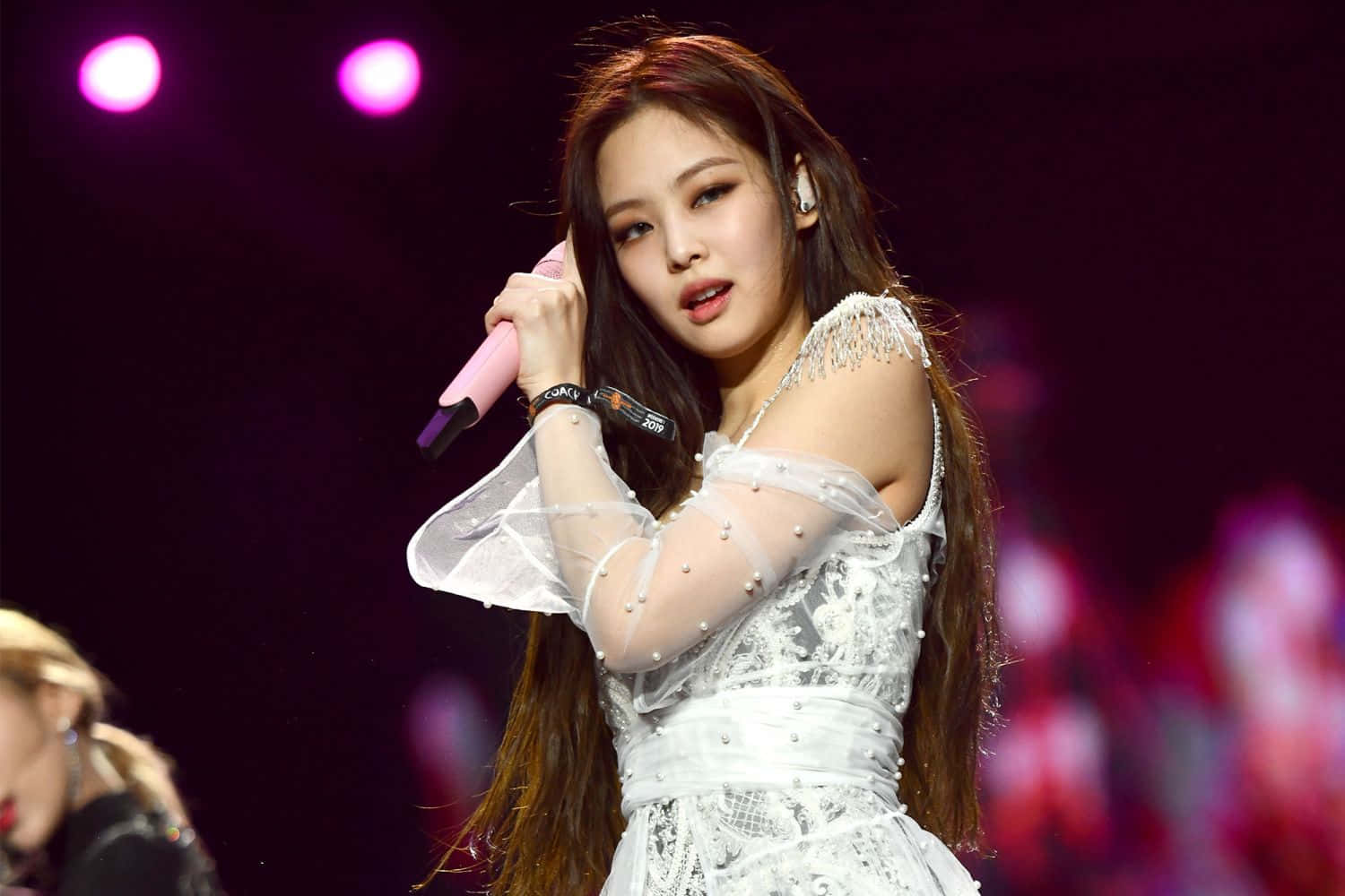 BLACKPINK Jennie Kim演唱會現場照片 壁紙 — 一位身穿白色蕾絲連衣裙的女歌手在粉紅色聚光燈下拿著粉紅色麥克風在舞台上表演。