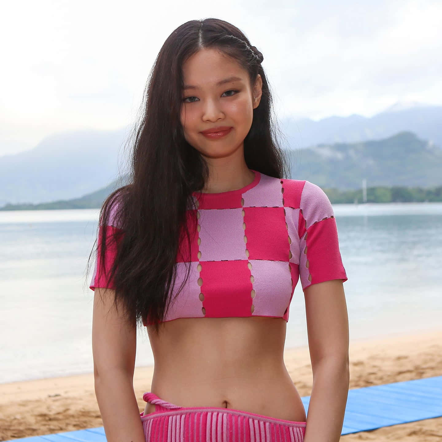 BLACKPINK Jennie Kim 時尚高質感照 壁紙 — 一位年輕女性身穿鮮豔的粉紅色格紋短版上衣，站在沙灘上，背景是平靜的海水和遠處的山脈，天空多雲。