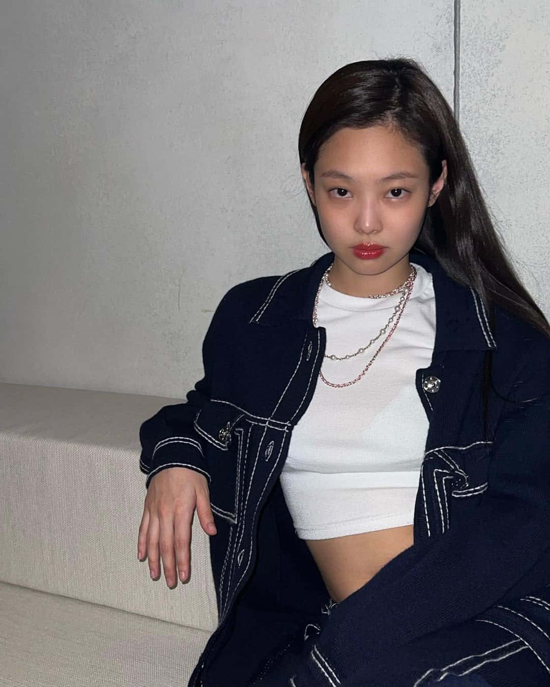 BLACKPINK Jennie身穿黑色牛仔外套照片 壁紙 — 一位長黑髮的年輕女性坐在米色沙發上，身穿白色車縫線的深藍色外套，內搭白色短版上衣，佩戴多層項鍊。