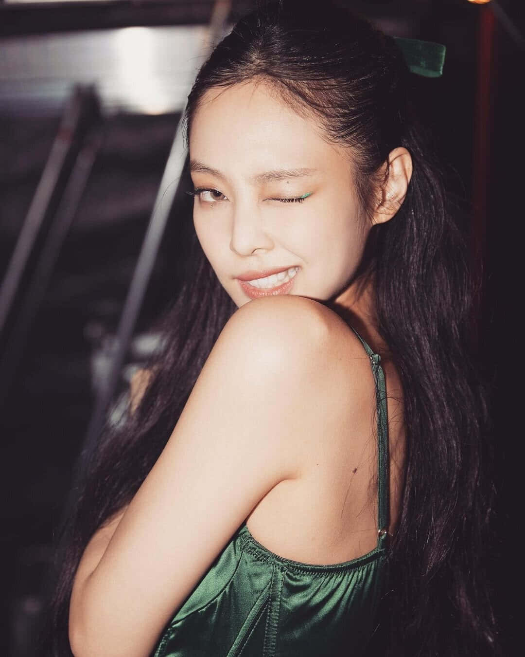 BLACKPINK Jennie Kim眨眼左眼照片 壁紙 — 一位長黑髮、綠色眼影的女性，在昏暗的室內環境中穿著閃亮的綠色連衣裙，頑皮地微笑。