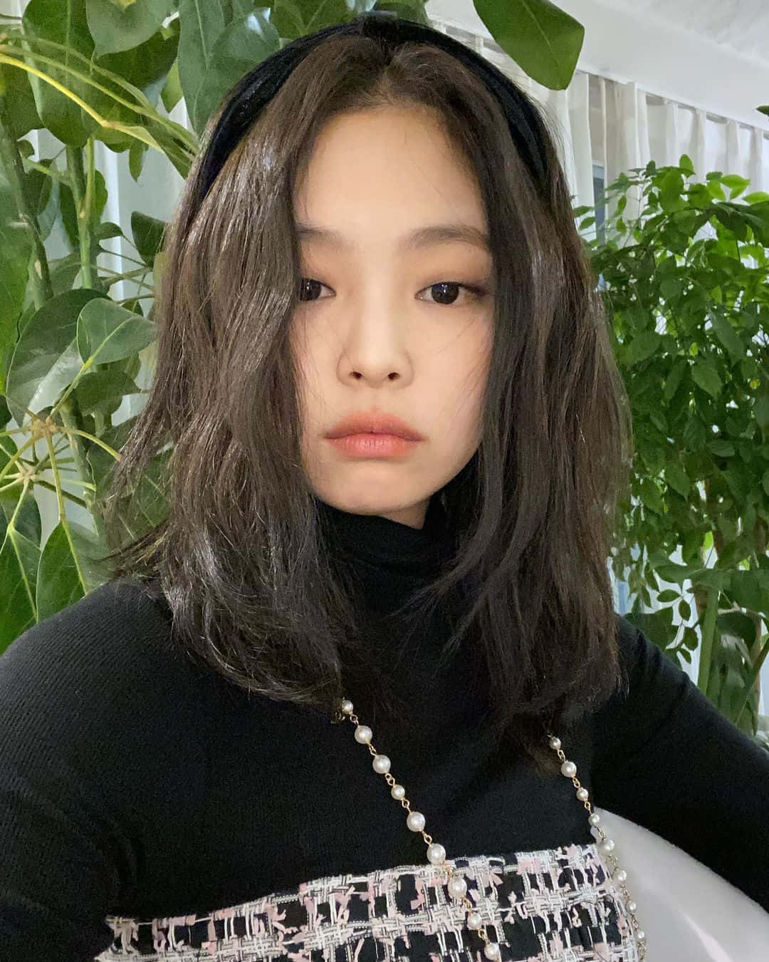 BLACKPINK Jennie Kim與植物合照自拍 壁紙 — 一位年輕女性，深色波浪長髮配黑色髮帶，身穿黑色高領衫，手持珍珠裝飾手袋，在茂密綠葉前溫柔凝視。