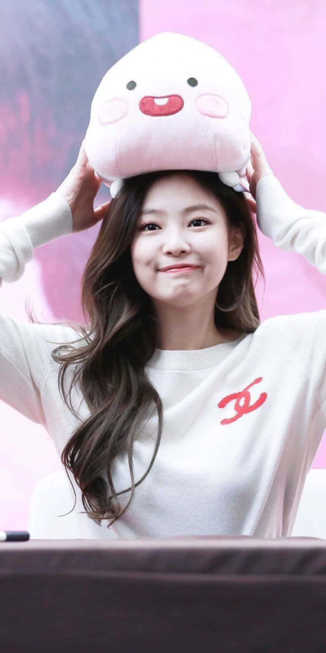 BLACKPINK Jennie Kim與可愛毛絨玩具合照 壁紙 — 一位微笑的年輕女性，穿著帶有紅色標誌的白色毛衣，頑皮地將一個粉紅色毛絨角色平衡在頭上。